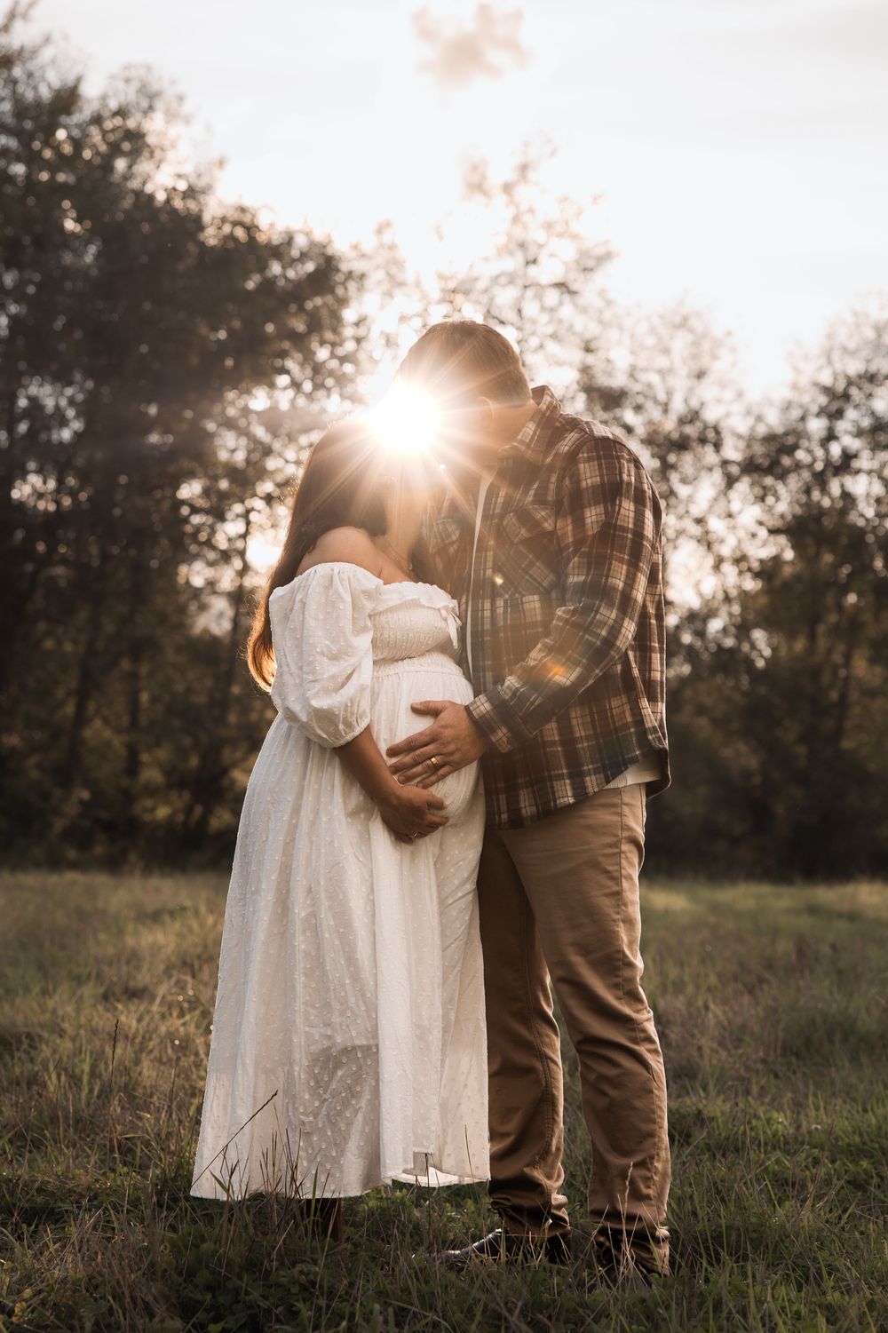Couple embracing at golden hour in a field, pregnant woman in white dress, man in plaid shirt, sun flare between them.