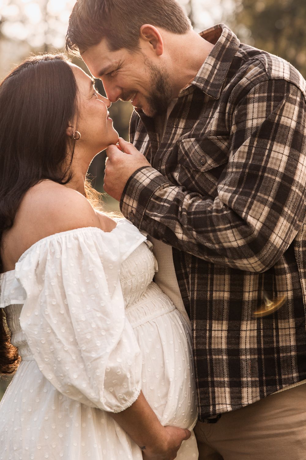Pregnant couple sharing a tender moment outdoors, woman in white off-shoulder dress, man in plaid shirt, golden hour light.