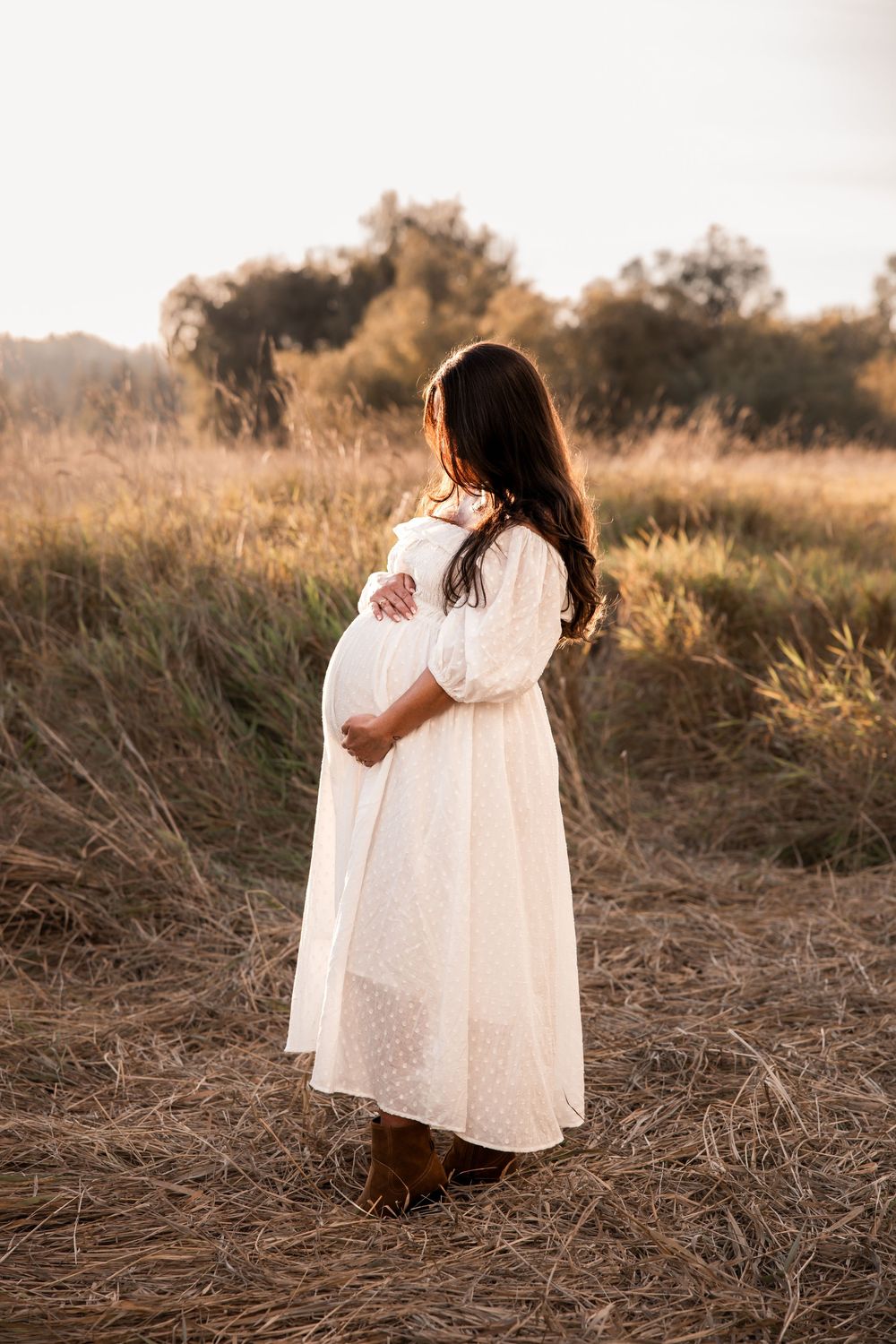 Pregnant woman in white dress holding baby bump in golden field at sunset.