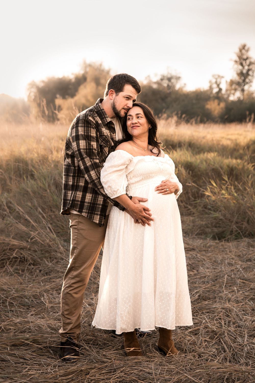 Couple embracing in a golden field, pregnant woman in white dress held by man in plaid shirt during maternity photoshoot.