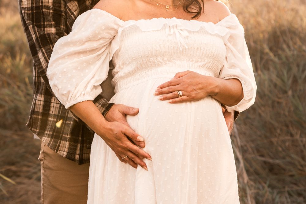 Couple in outdoor maternity photo shoot, woman in white off-shoulder dress cradling baby bump.