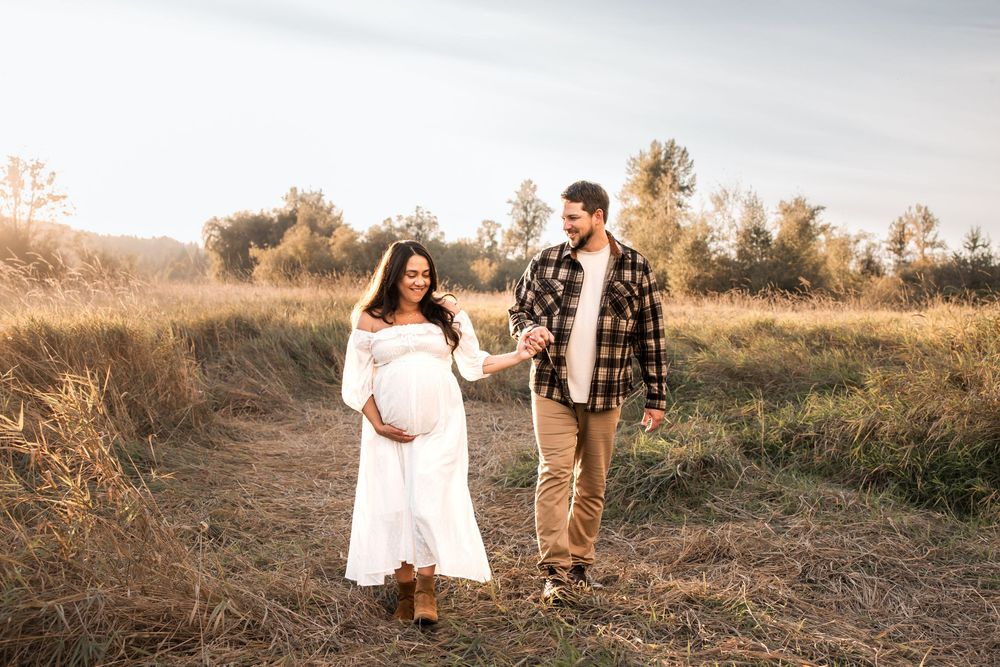 Pregnant couple holding hands walking through golden field at sunset, woman in white dress, man in plaid shirt.