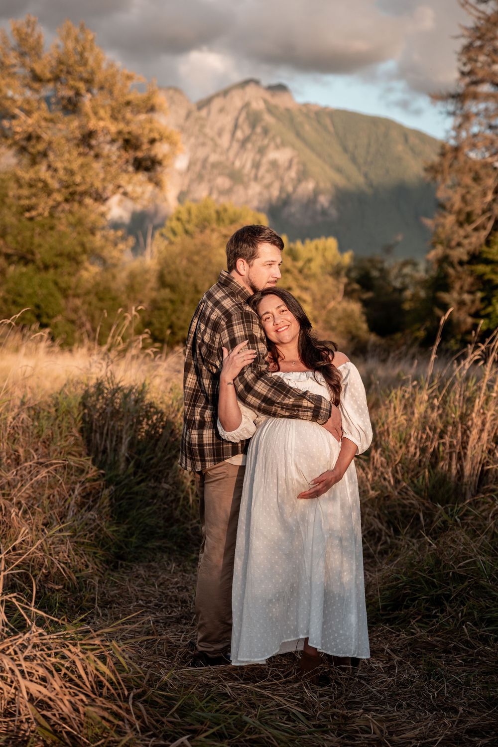 Couple shares tender maternity moment outdoors, woman in white dress cradling bump, mountain backdrop at golden hour.