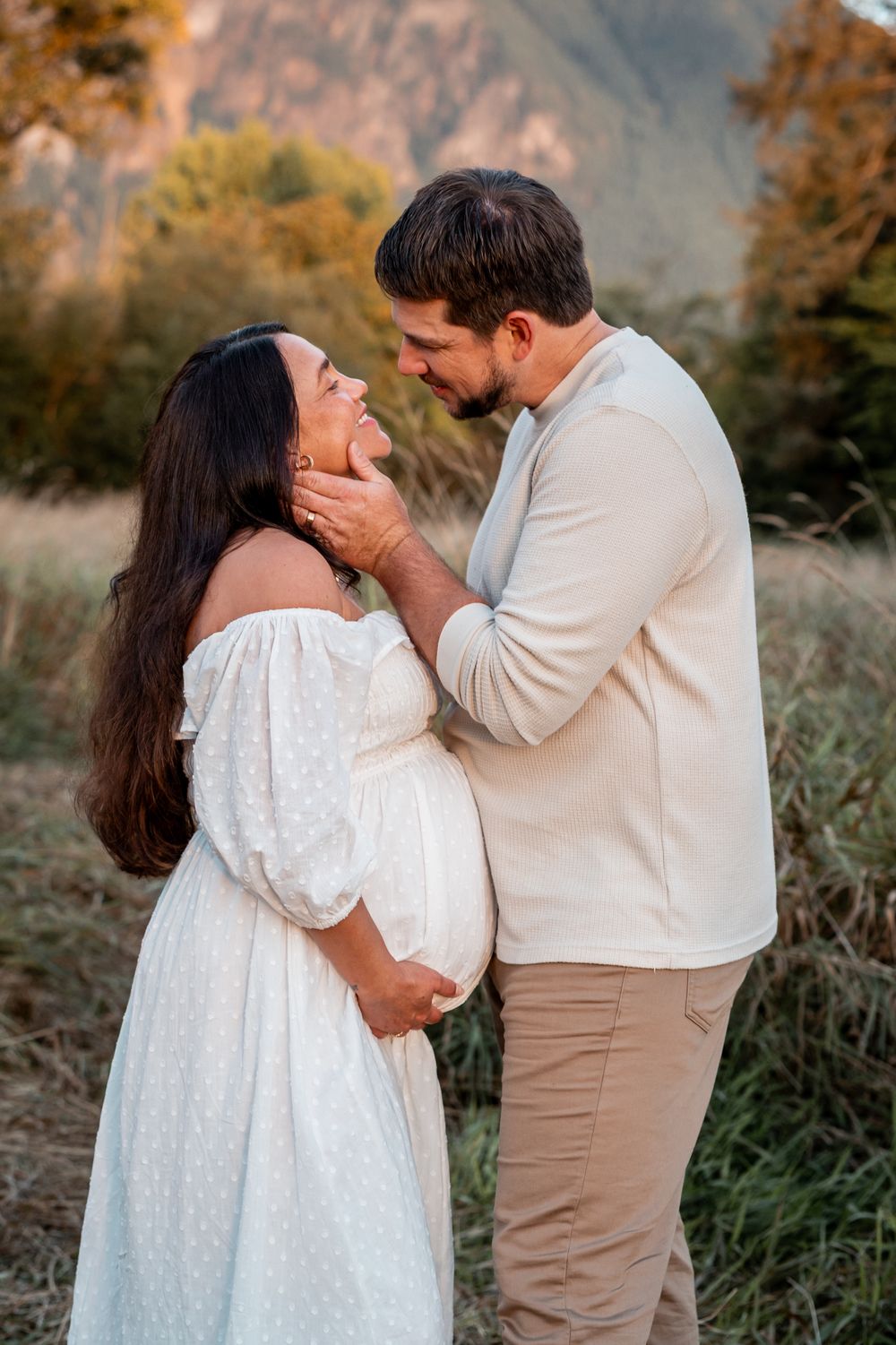 Couple sharing a tender moment outdoors, pregnant woman in white off-shoulder dress, man in beige, mountains behind.