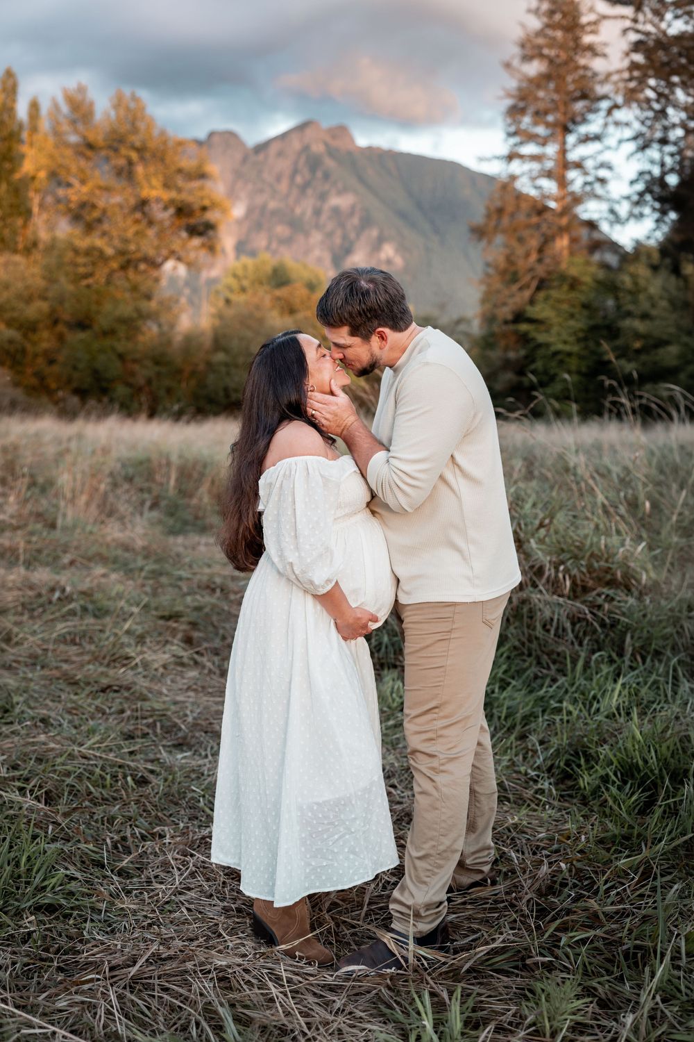 Couple sharing a tender kiss in a scenic mountain meadow, woman in white off-shoulder dress, golden autumn trees backdrop.