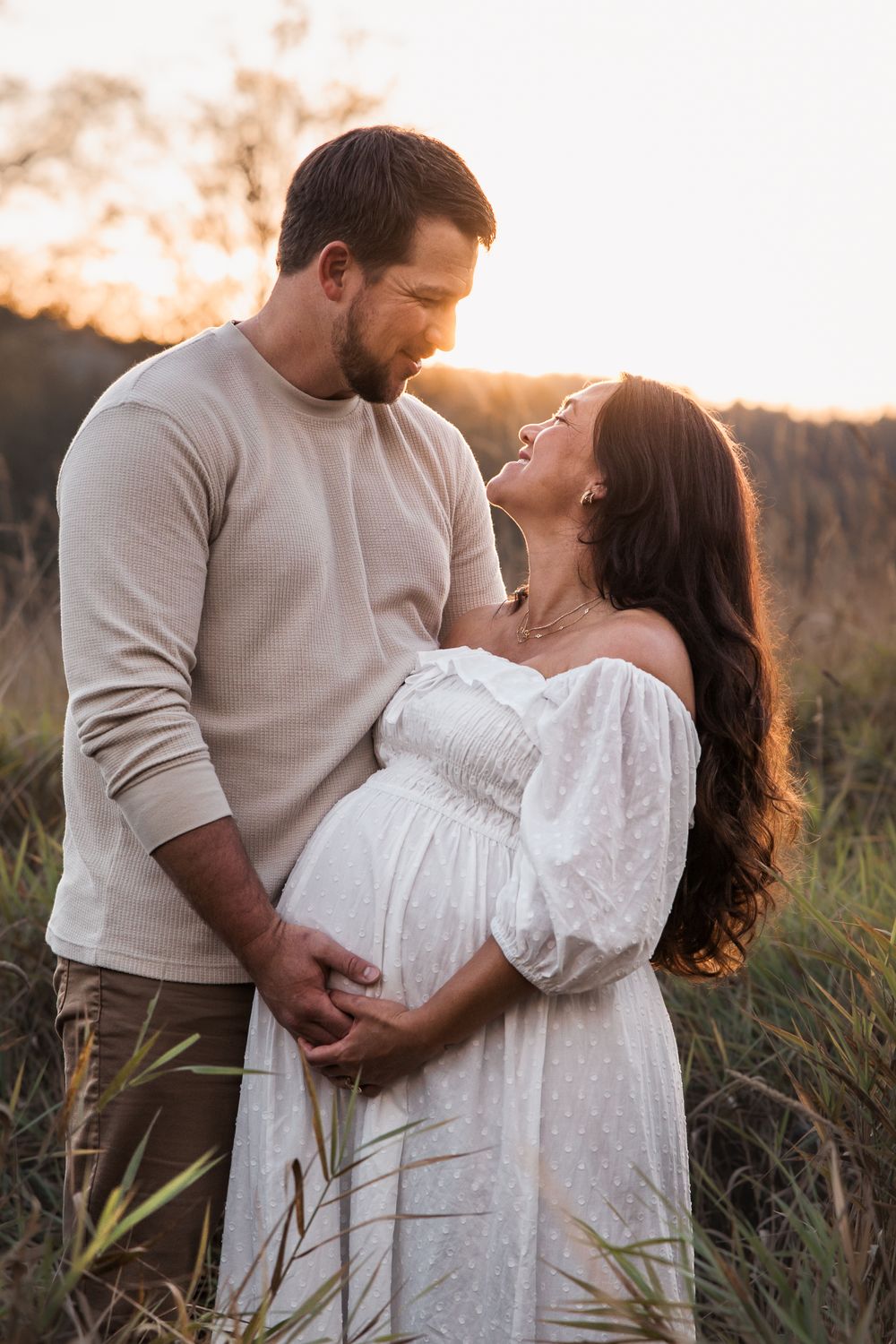 Couple embracing at golden hour sunset in a field, pregnant woman in white dress smiling up at man in beige sweater.