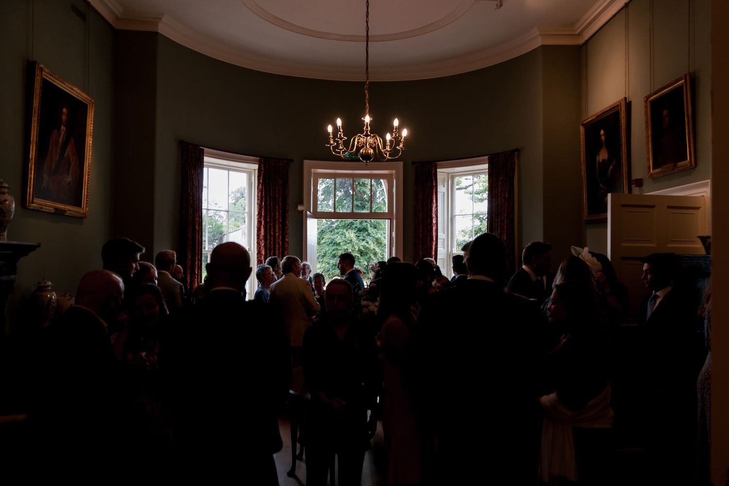 A dimly lit historic room with a chandelier and large windows shows a crowd of people gathered in silhouette.