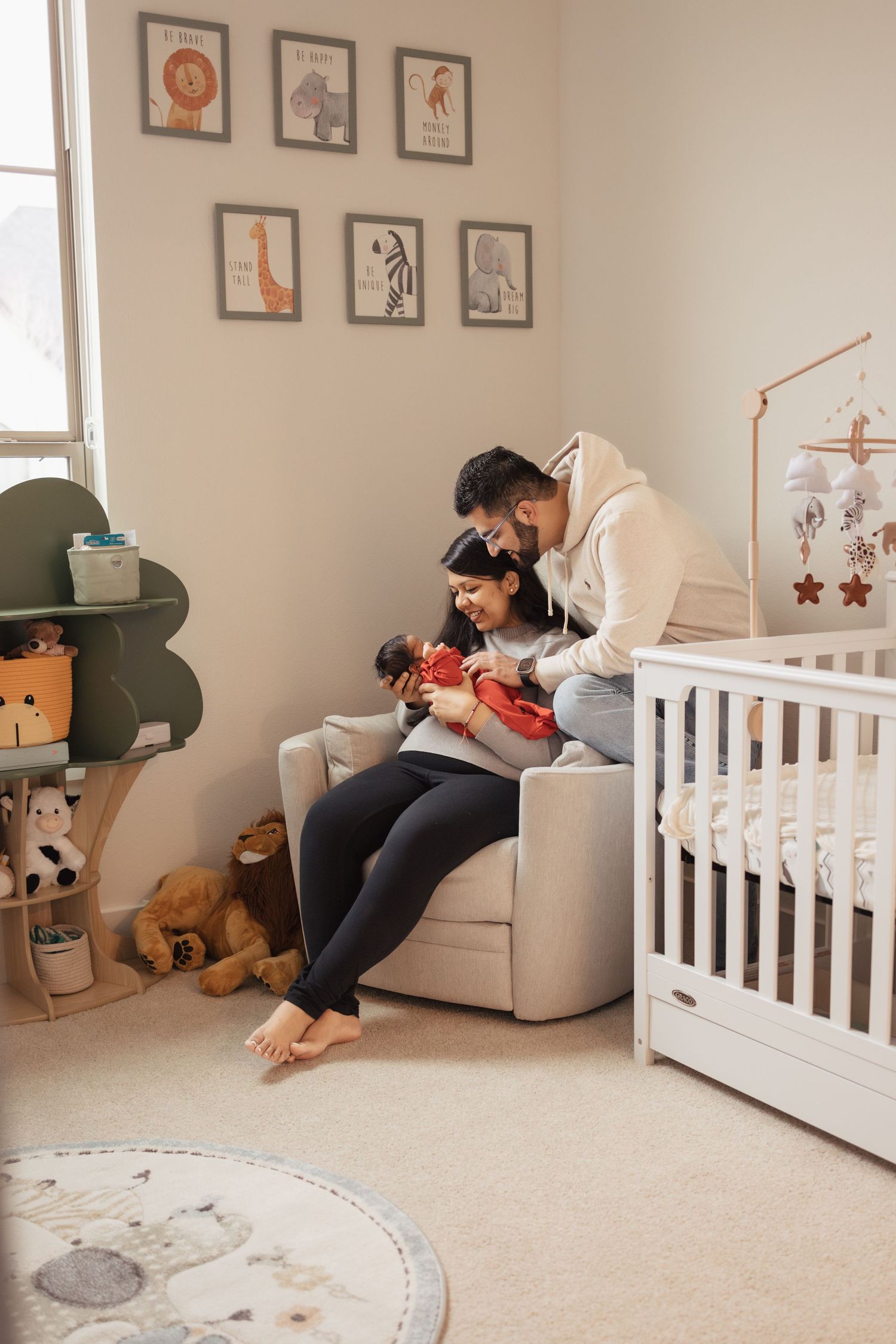 A cozy nursery room with framed art prints, stuffed animals, white crib, and comfortable armchair beside a window.