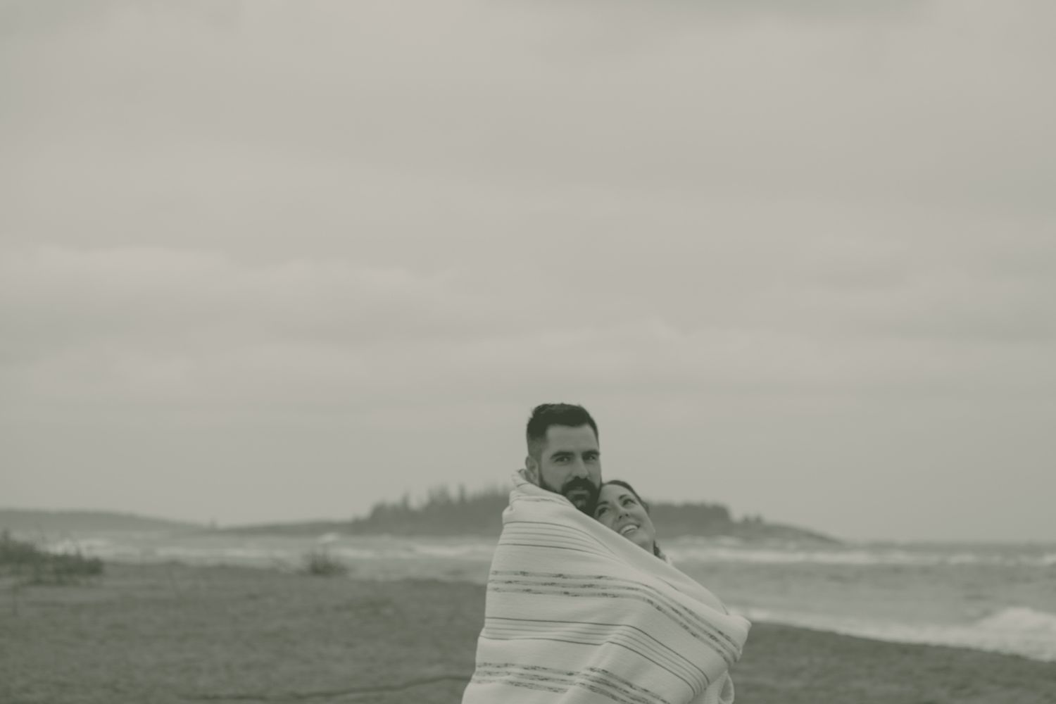 An engaged couple lays on a blanket smiling at each other