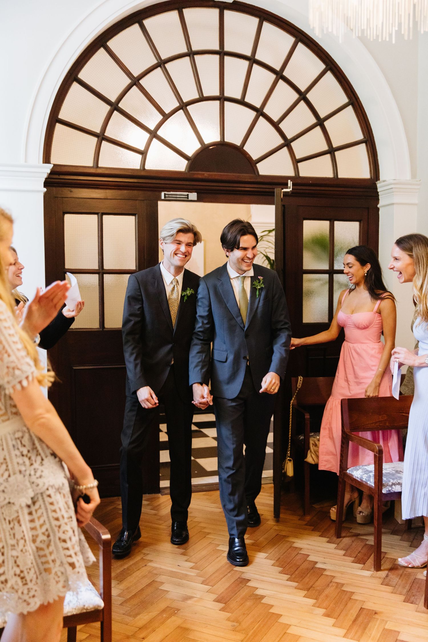 Wedding party members in formal attire walk through an elegant doorway with a sunburst transom window.