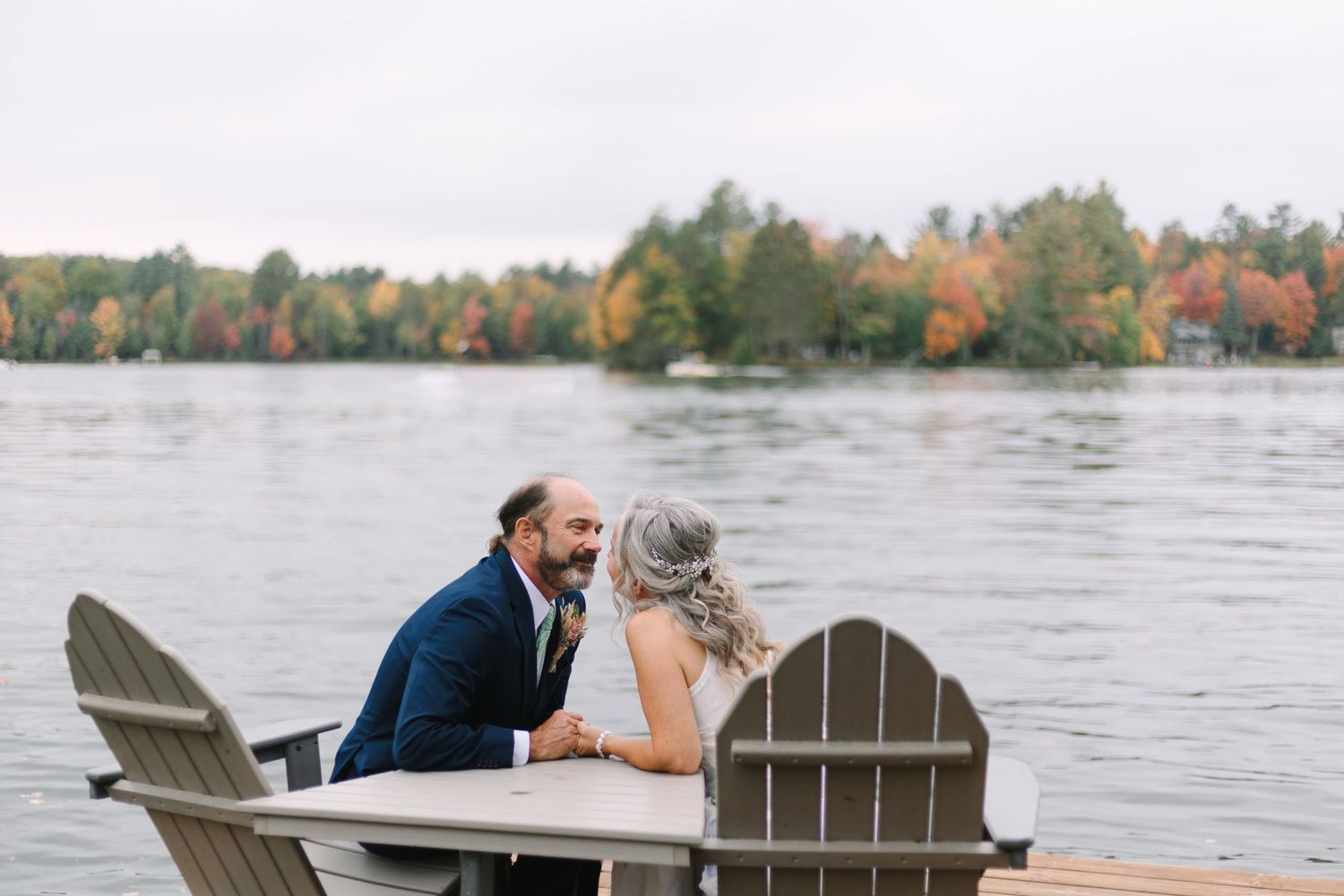 Two people seated in Adirondack chairs share an intimate moment on a dock by a lake with autumn trees in background.