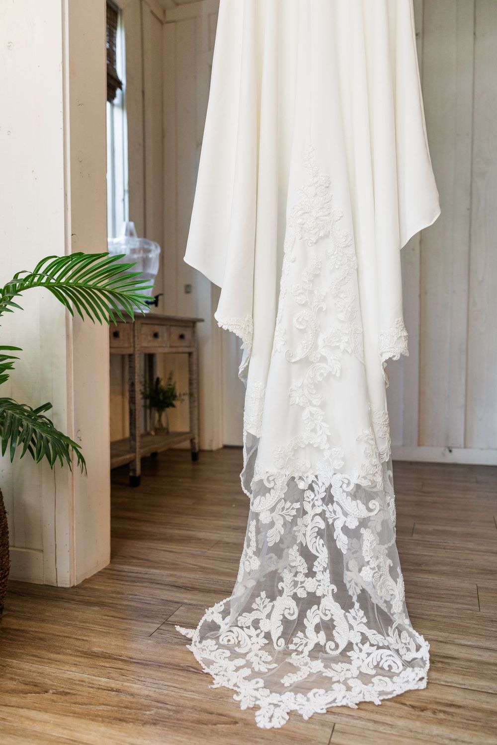 Elegant white wedding dress with lace trim hanging against white wall with hardwood floors.