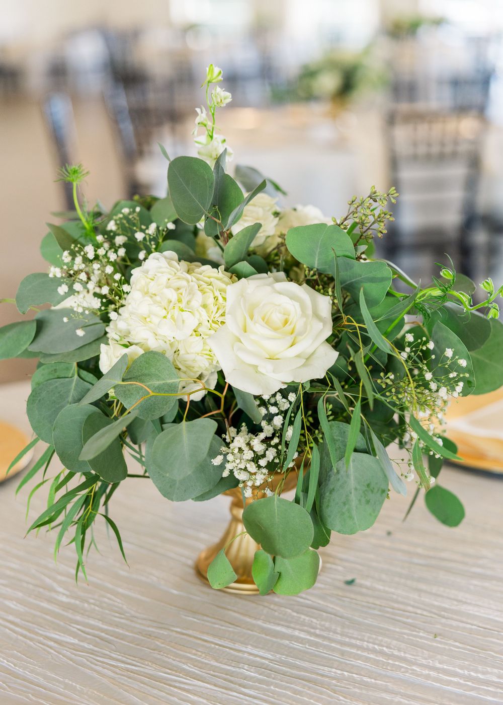 White roses and eucalyptus centerpiece arranged in gold vase on gray table.