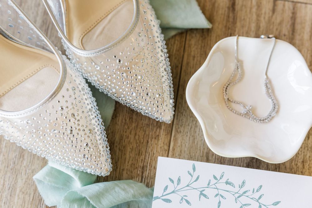 Wedding accessories including sparkly pointed shoes and delicate necklace on wooden surface.