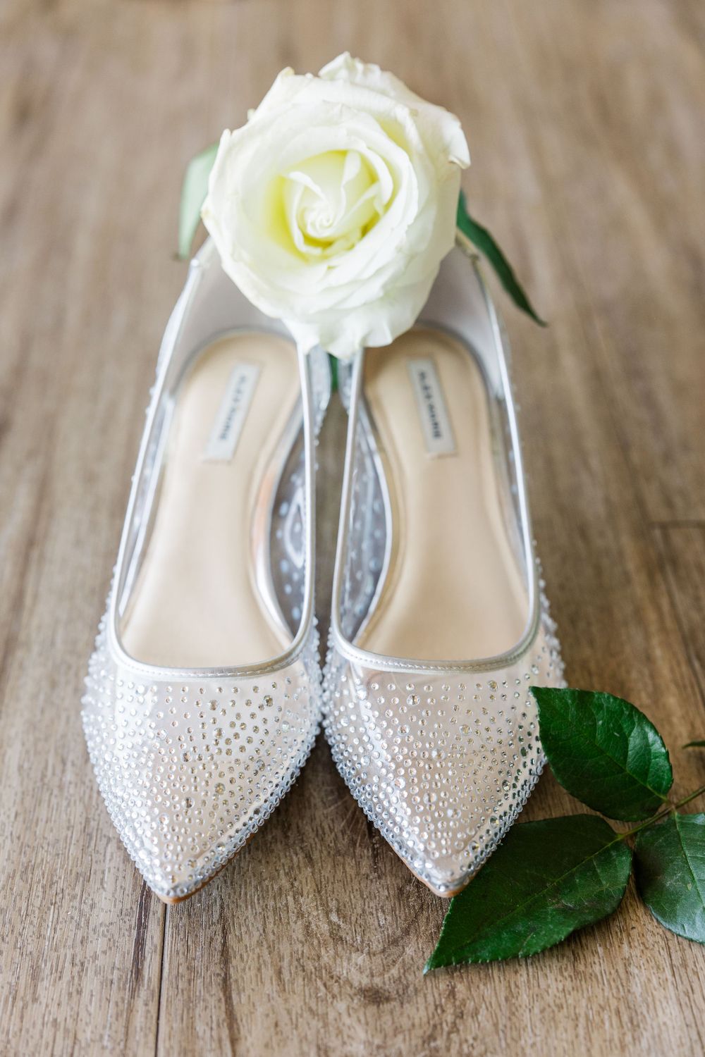 Elegant white rhinestone wedding flats with a single white rose on a wooden surface.
