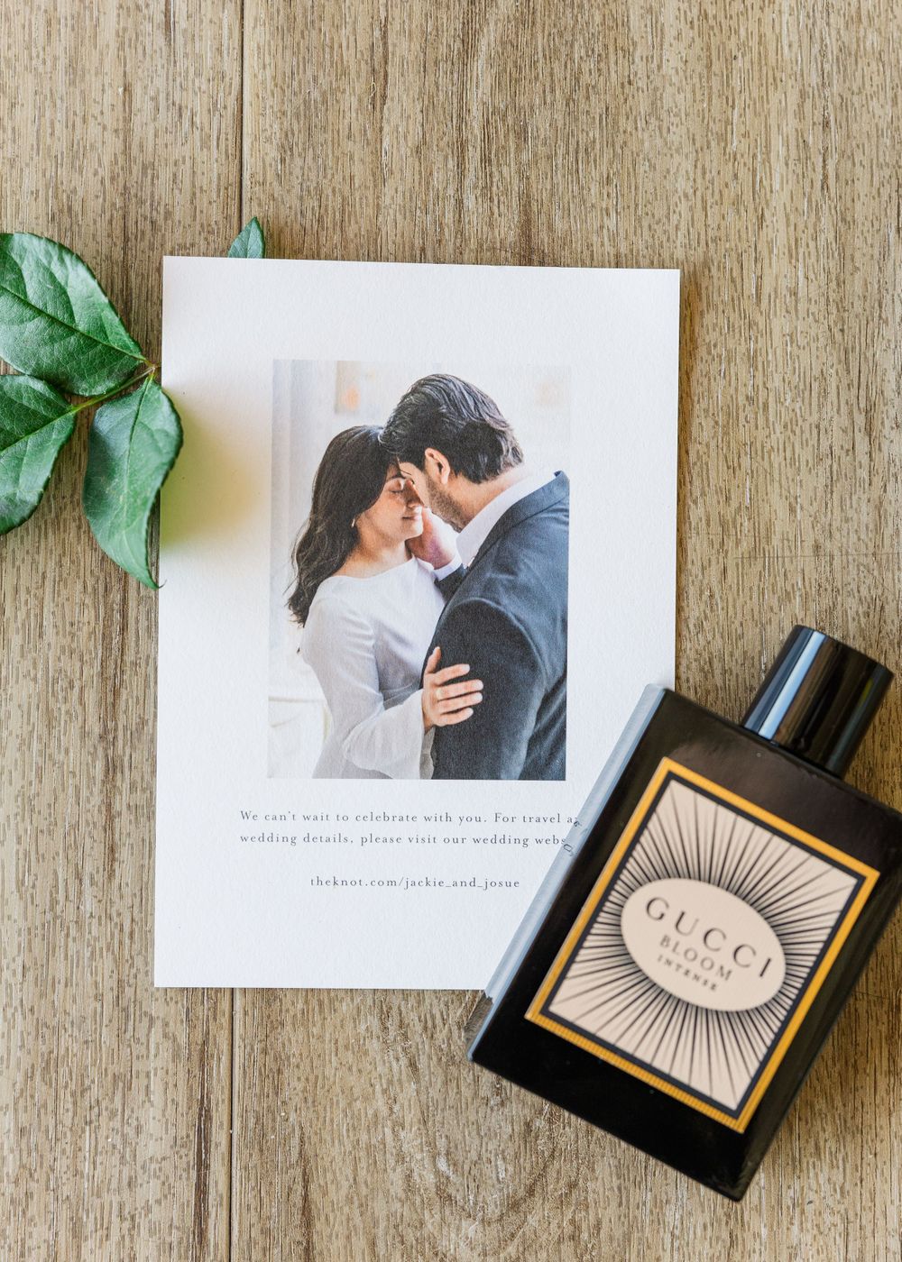 Wedding invitation and Gucci perfume bottle arranged on wooden surface with green leaf accent.