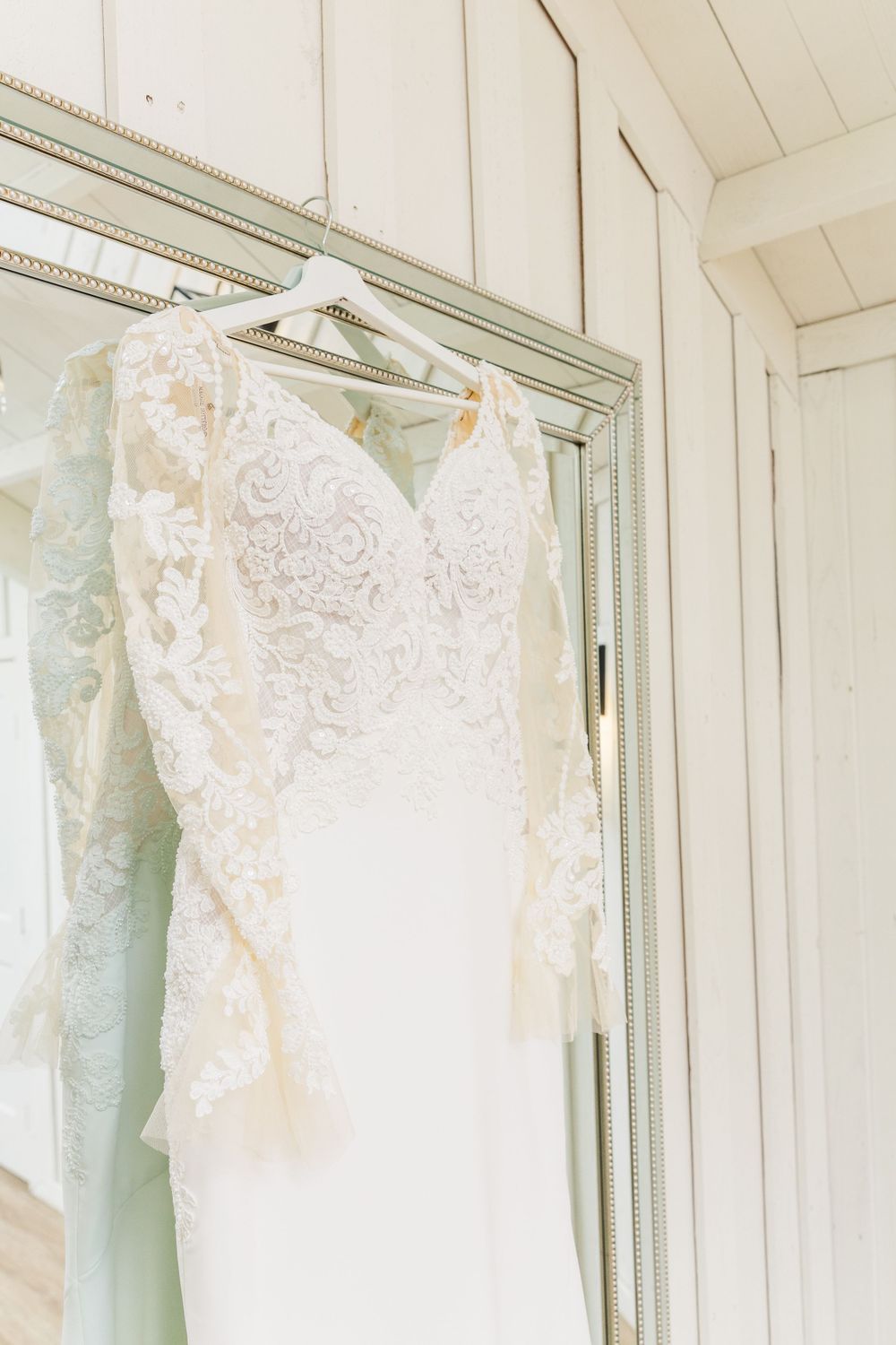 An elegant white lace wedding dress hangs on a vintage mirror in a light-filled room.