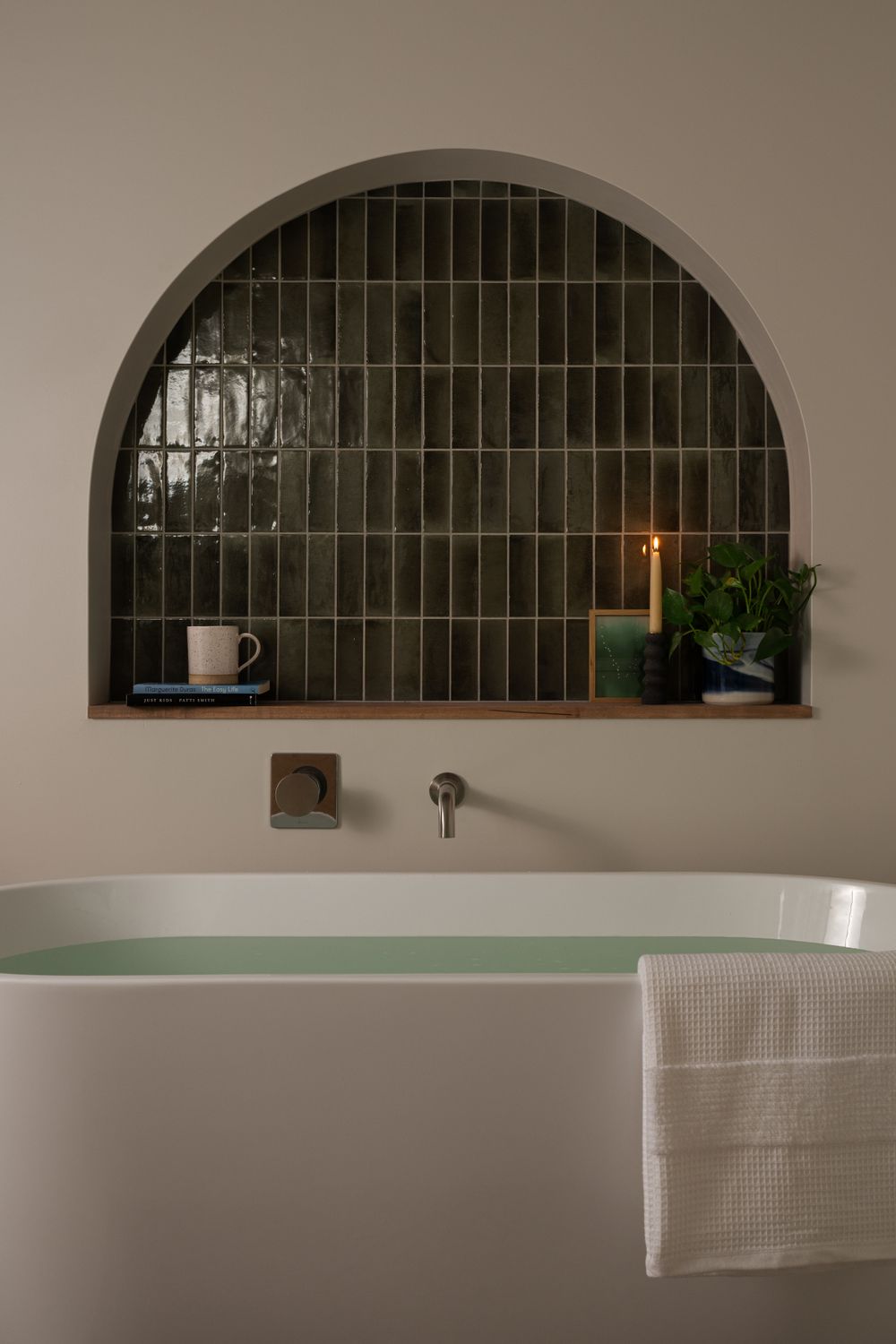 Arched wall niche with green tile backsplash above white freestanding bathtub, featuring decorative plants and a coffee cup.