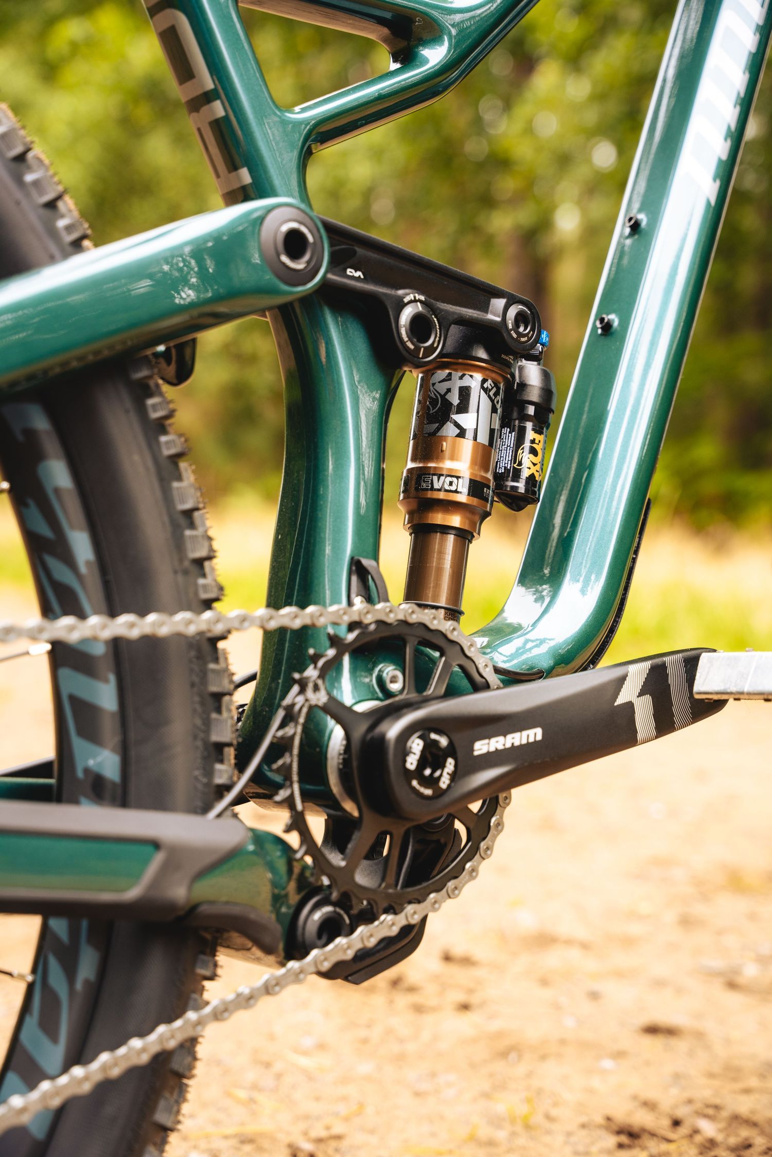 Close-up detail of a green mountain bike's rear suspension and drivetrain components on a dirt trail.