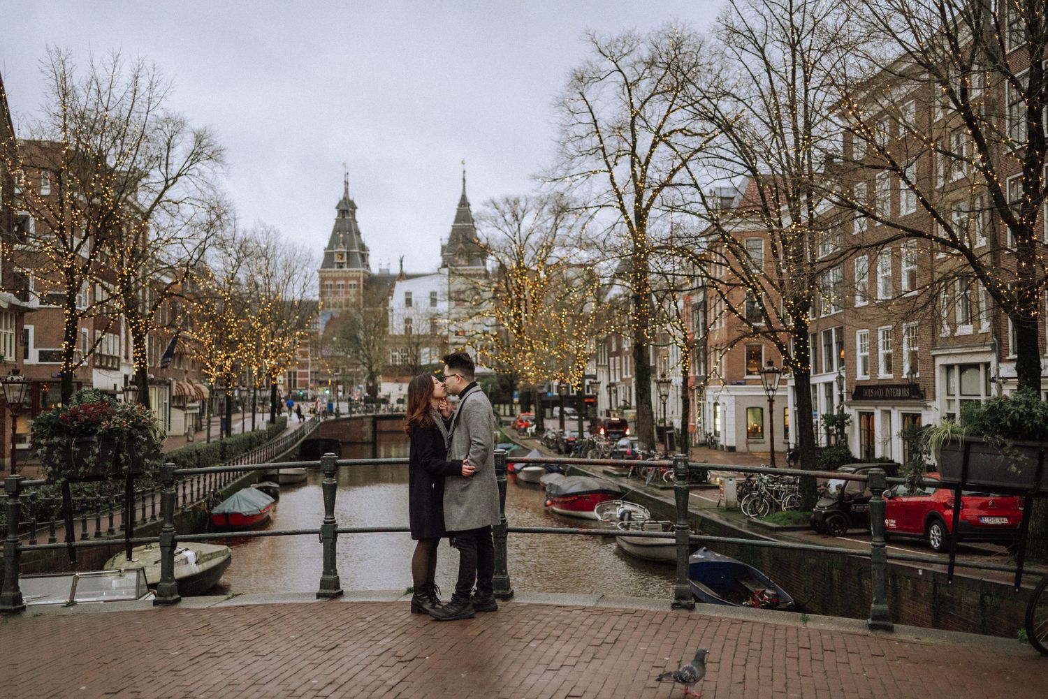 Couple standing in the middle of a bridge over a canal in central Amsterdam, with the Rijksmuseum towers in the background.