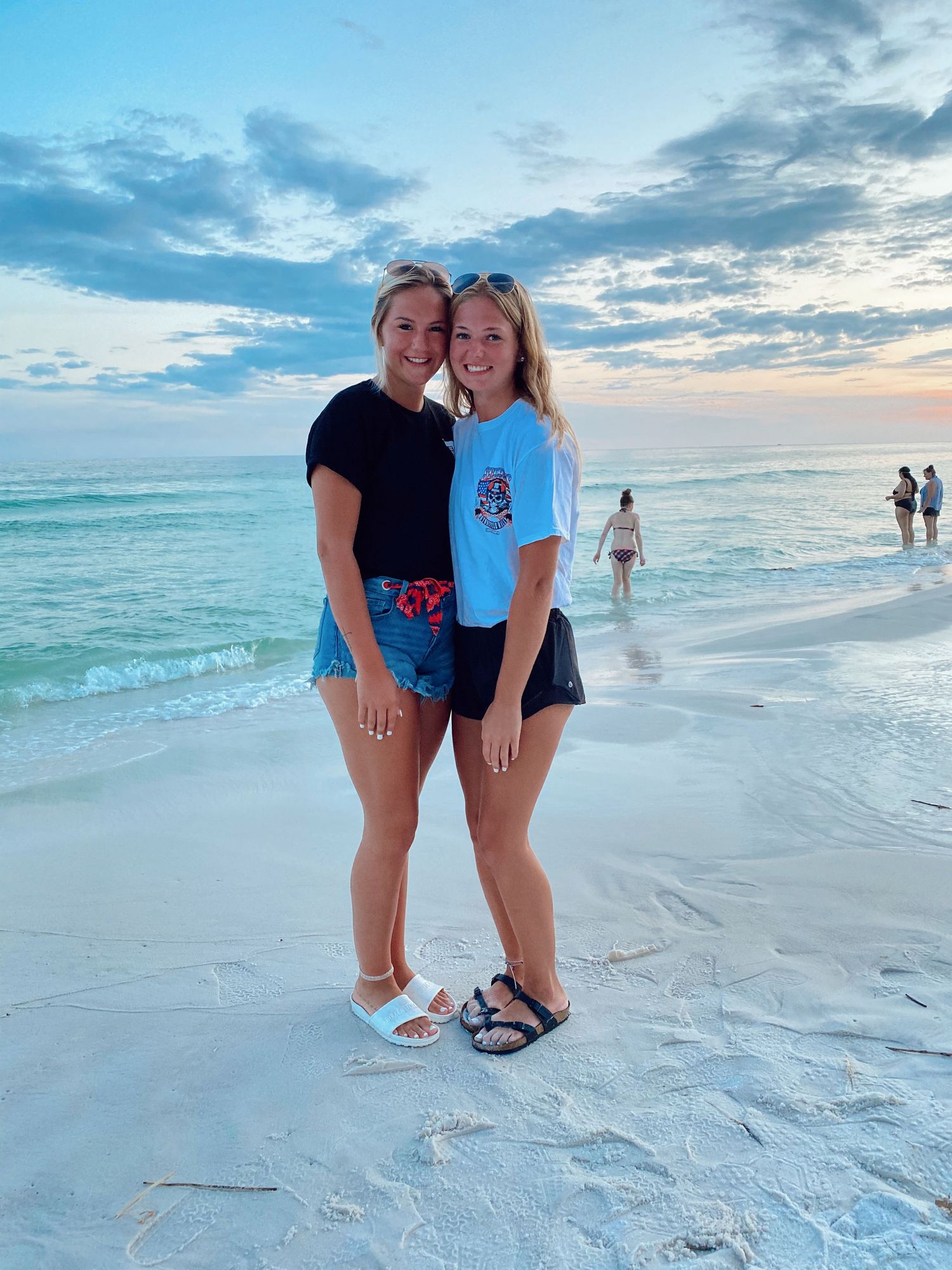 Two people in casual beach attire pose together on a white sandy beach at sunset with turquoise ocean waves behind them.