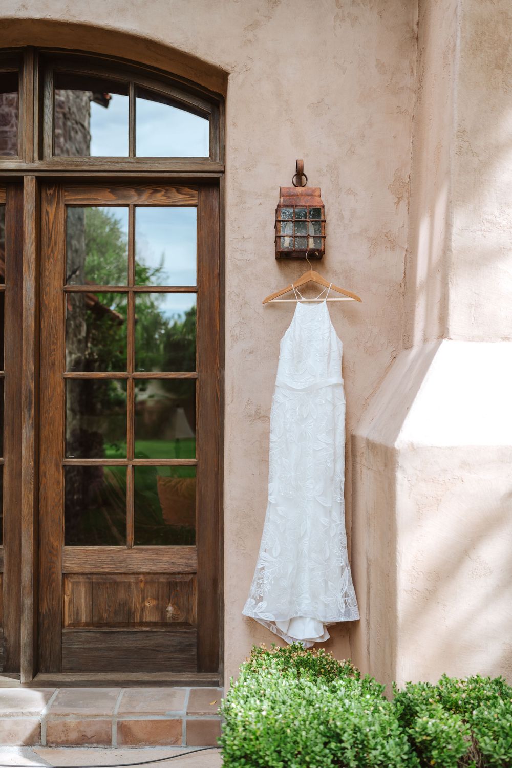 A white lace wedding dress hangs on a rustic adobe wall with wooden windows and green plants in a romantic setting.
