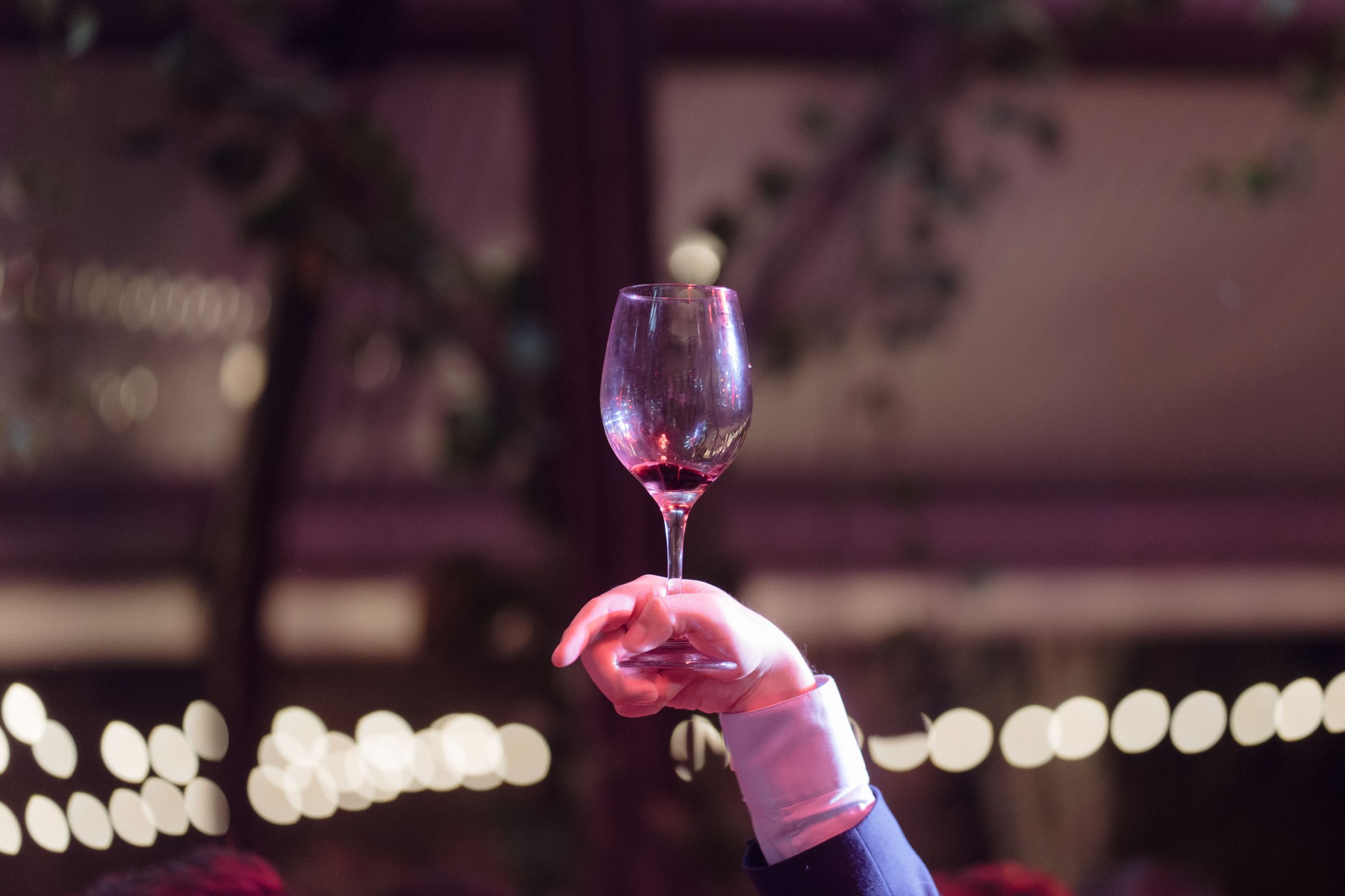 Hand holding glass of red wine with bokeh lights in background.