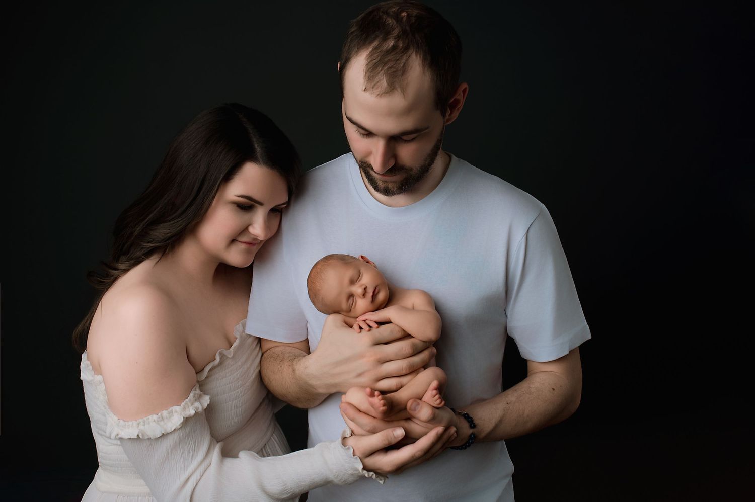 Parents lovingly hold and gaze at their newborn baby against dark backdrop during intimate portrait session.