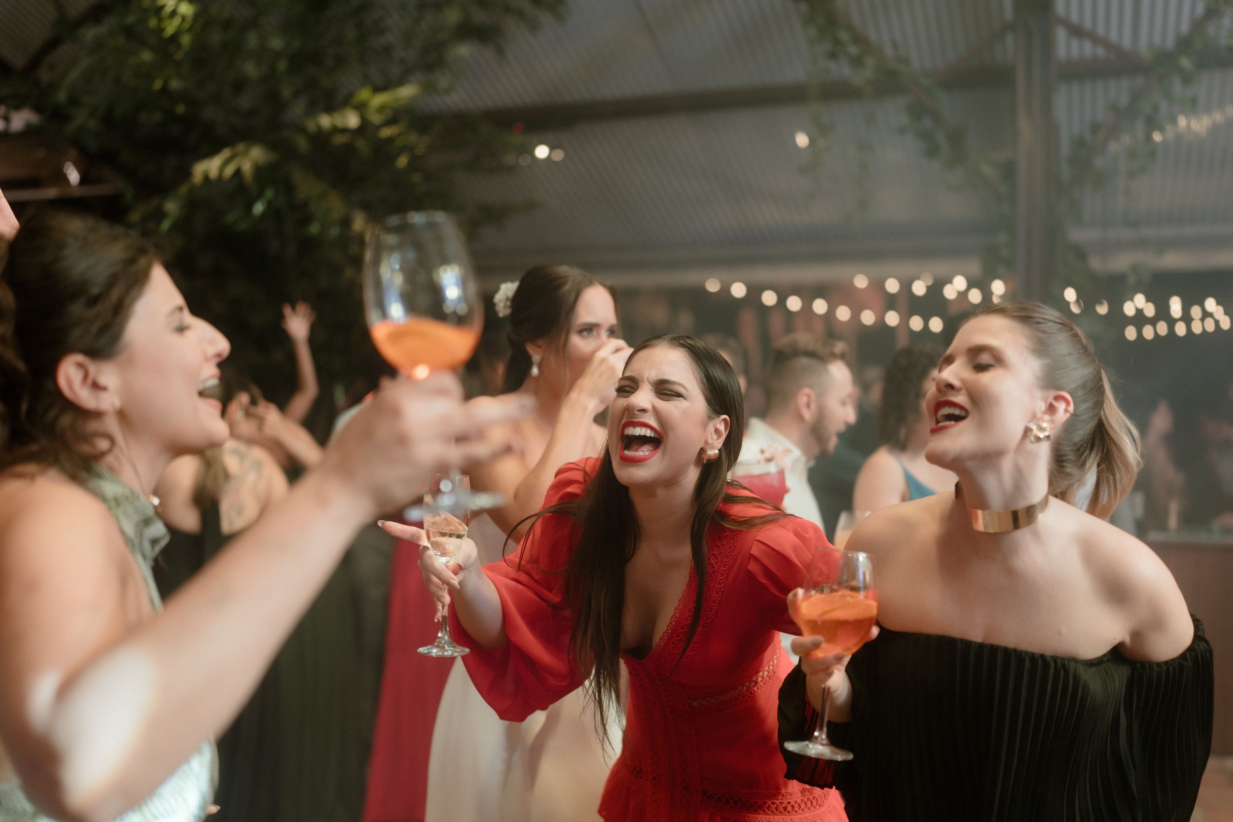 Party guests laughing and toasting with drinks at an outdoor celebration under string lights.