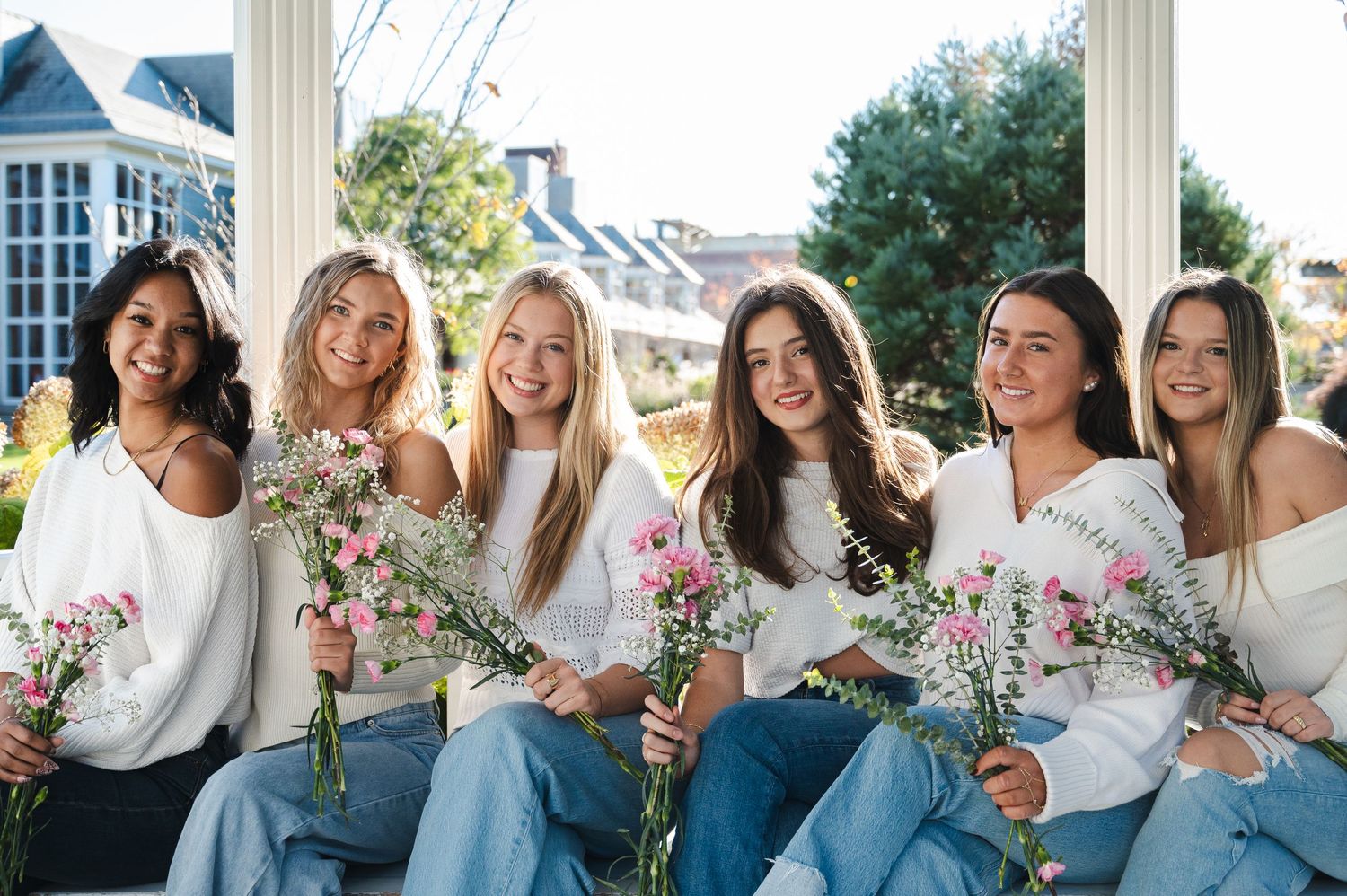Group photo of six women in white tops sitting together with flowers outdoors.