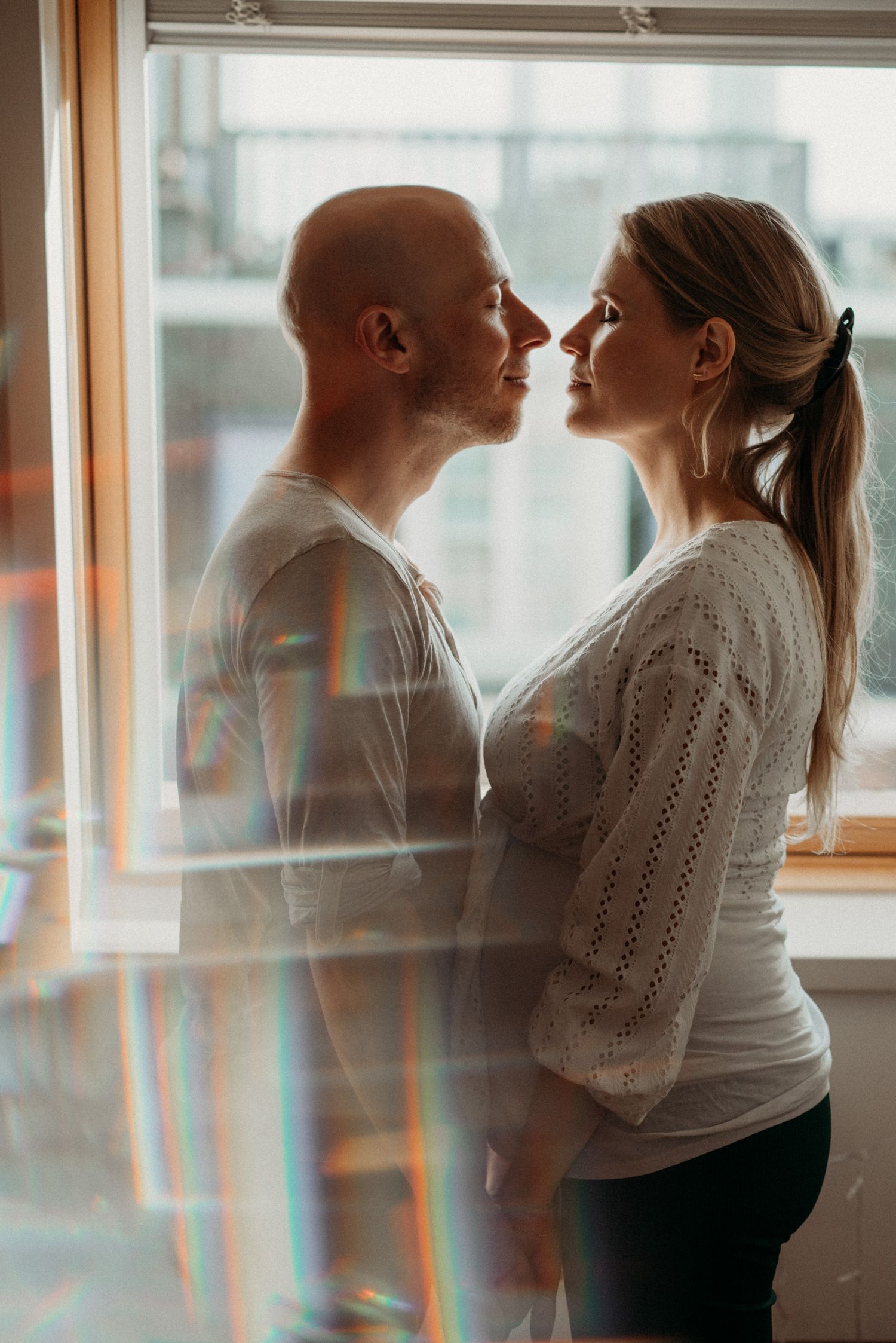 Romantic couple’s maternity photo in front of a window, cozy home session.