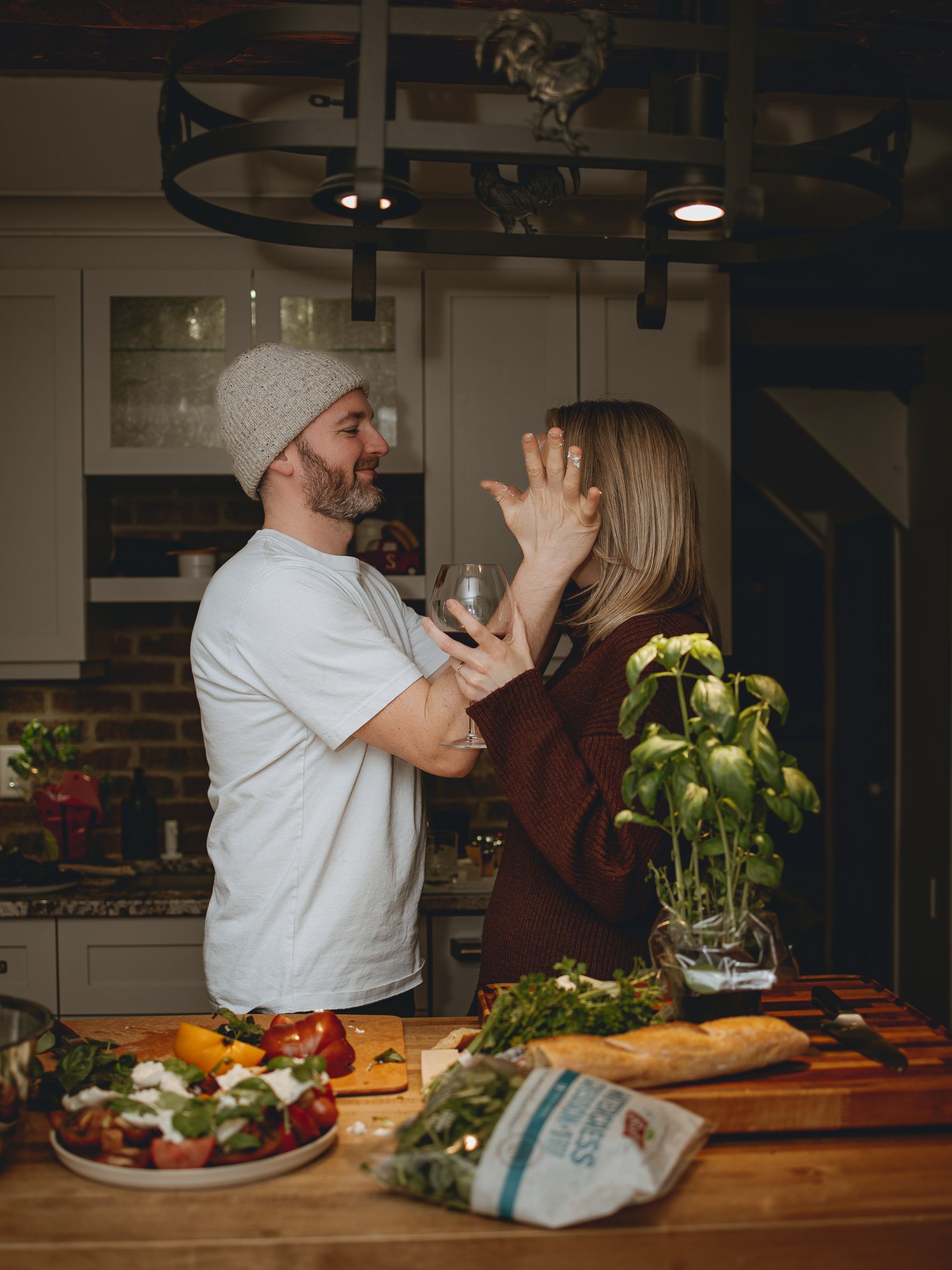 A playful cooking scene shows a couple laughing together in a modern kitchen with ingredients spread out.