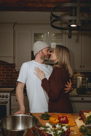 A couple shares an intimate moment while cooking together in a kitchen with fresh vegetables on the counter.
