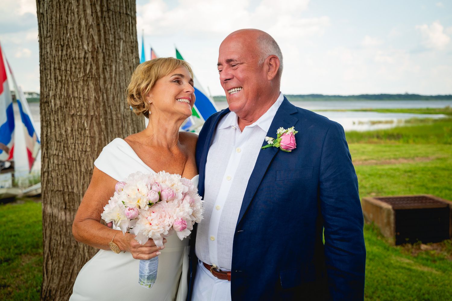 A happy couple shares a joyful moment outdoors by a tree with water in the background on their wedding day.