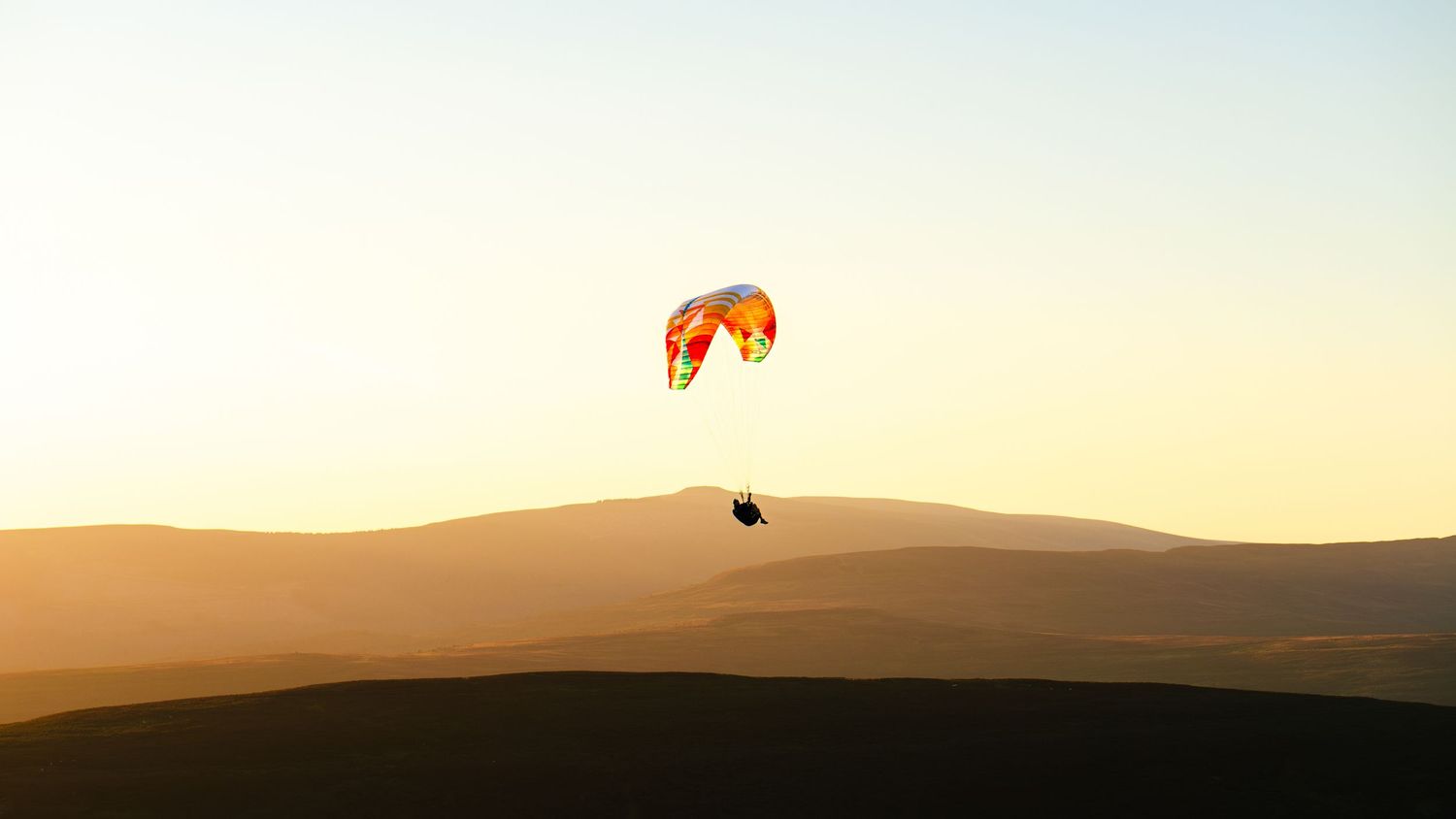 A paraglider soars above golden sand dunes at sunset with an orange wing against the warm sky.