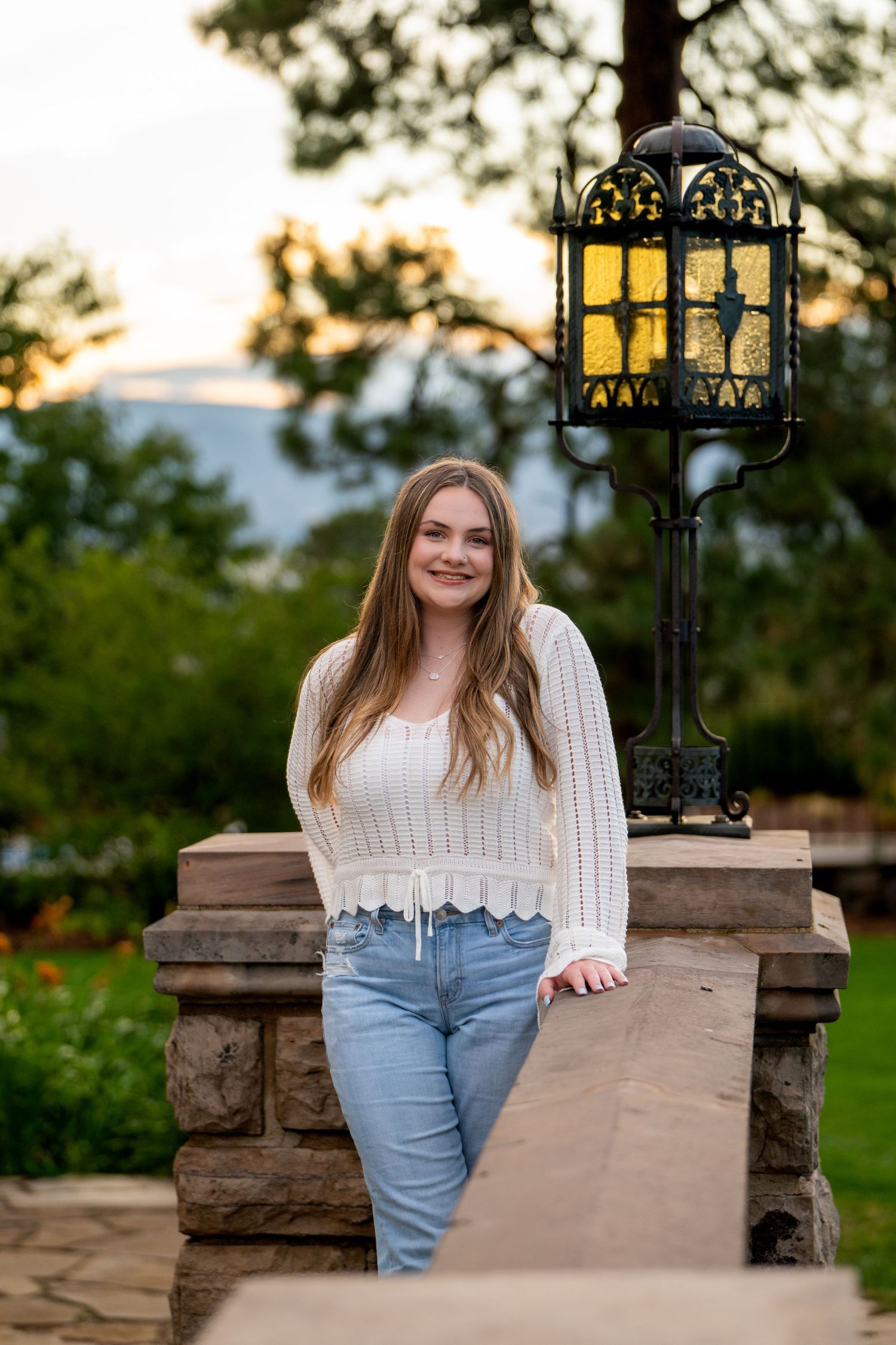 A person in a white knit sweater and jeans stands by a decorative iron lamppost at sunset in an outdoor garden setting.