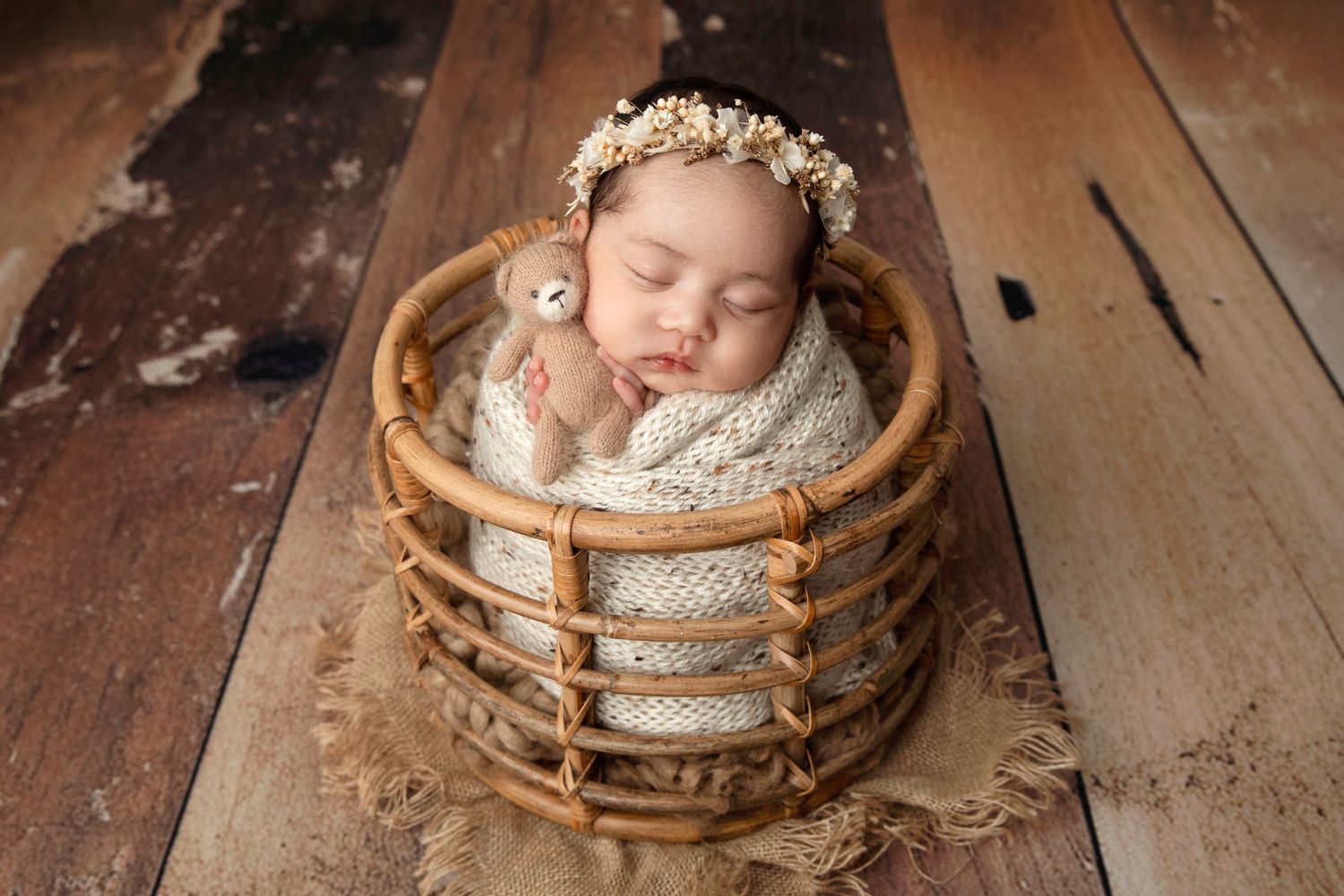 Sleeping newborn with flower crown nestled in round wicker basket on wooden floor.