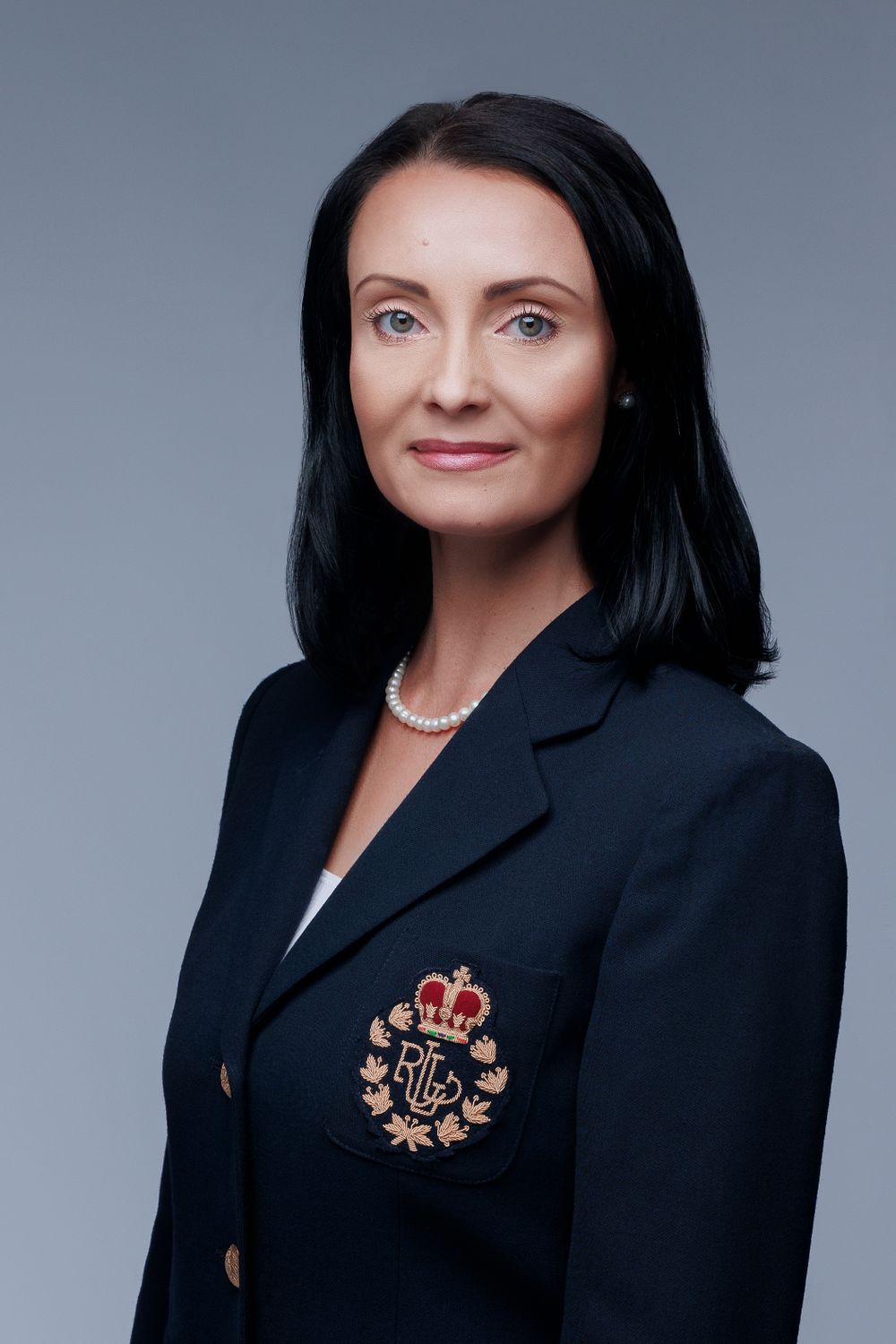 Professional portrait of woman in navy blazer with embroidered crest against gray backdrop.