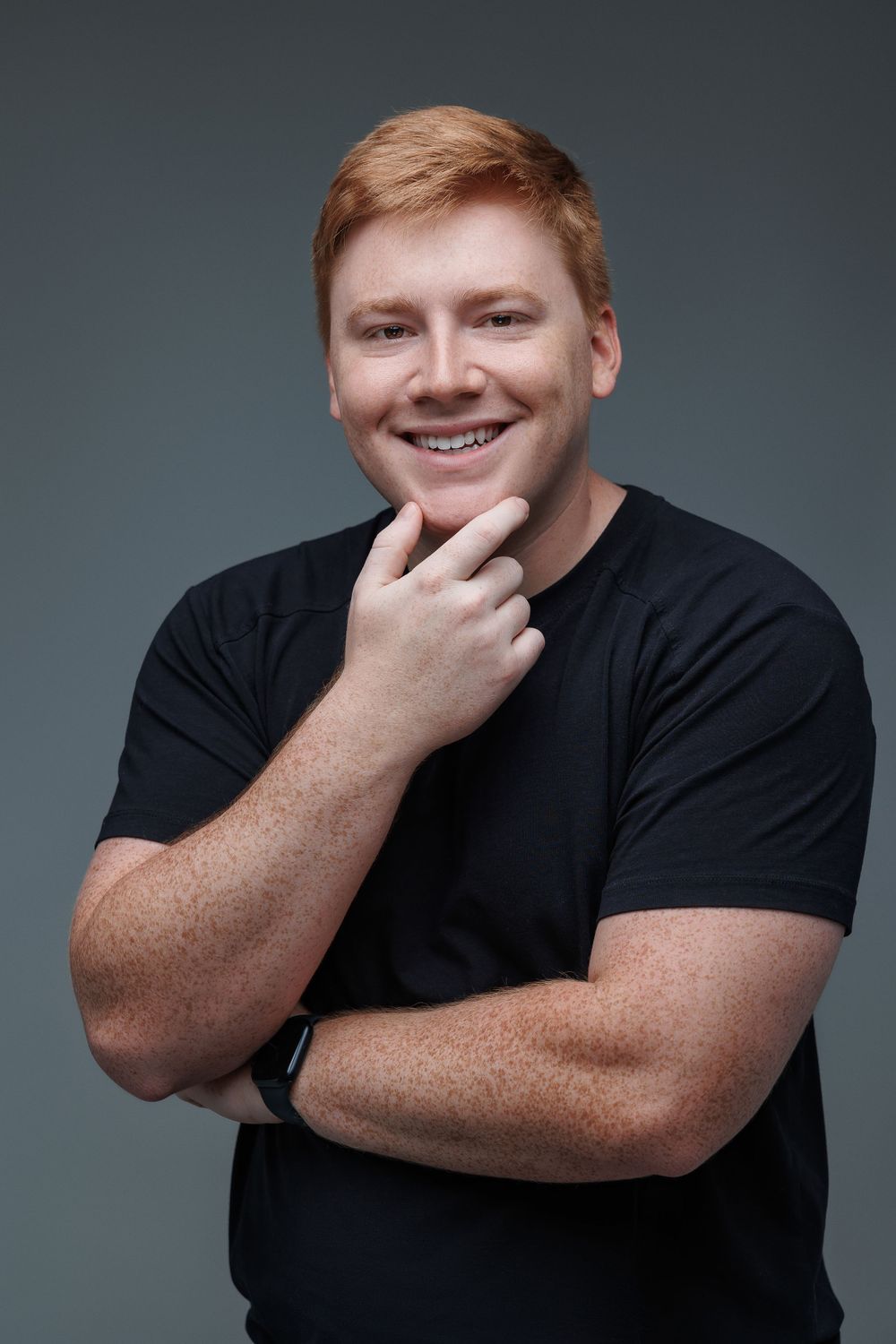 Portrait of man in black t-shirt with hand on chin against gray background.
