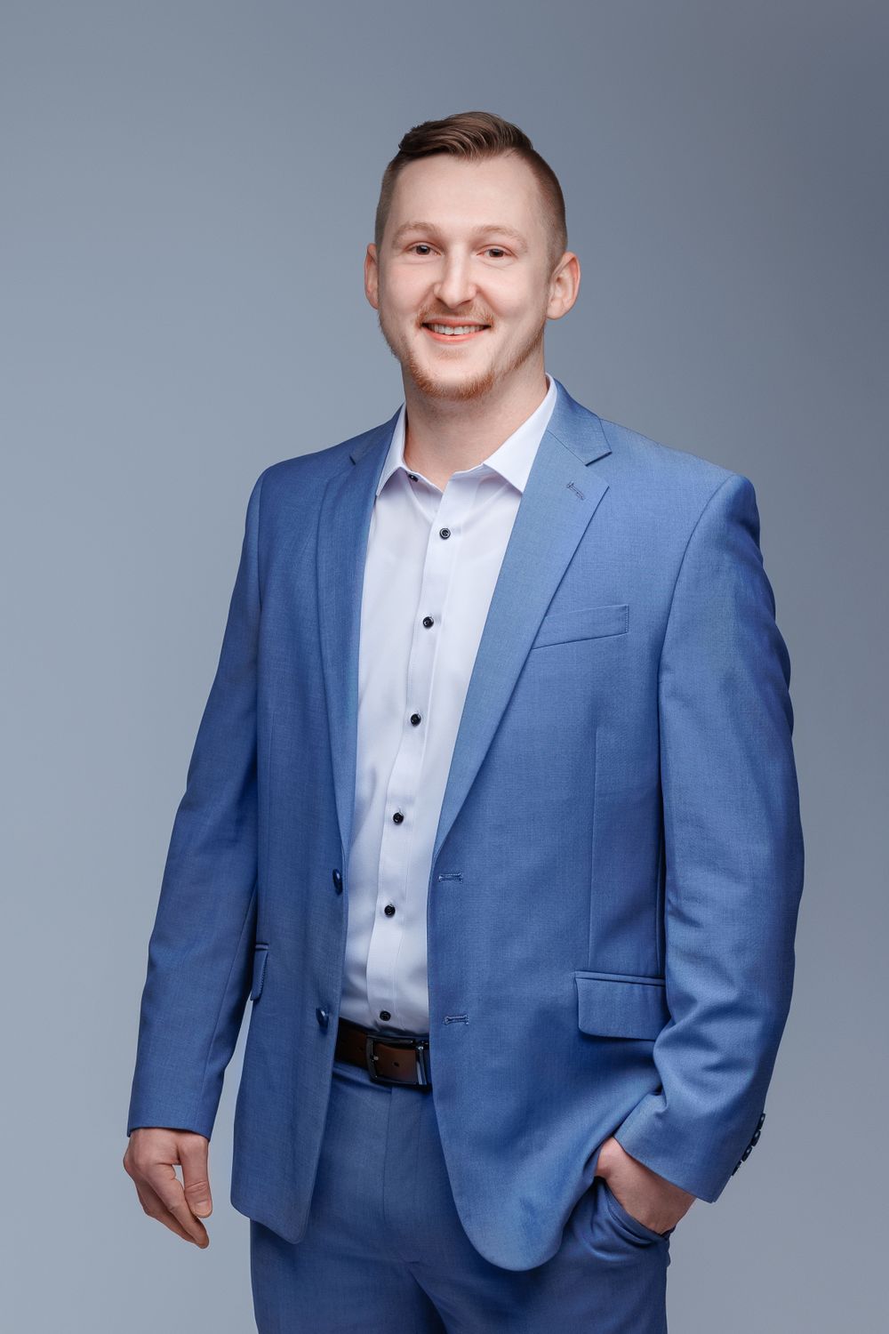 Professional headshot of man in blue suit with confident smile against grey backdrop.