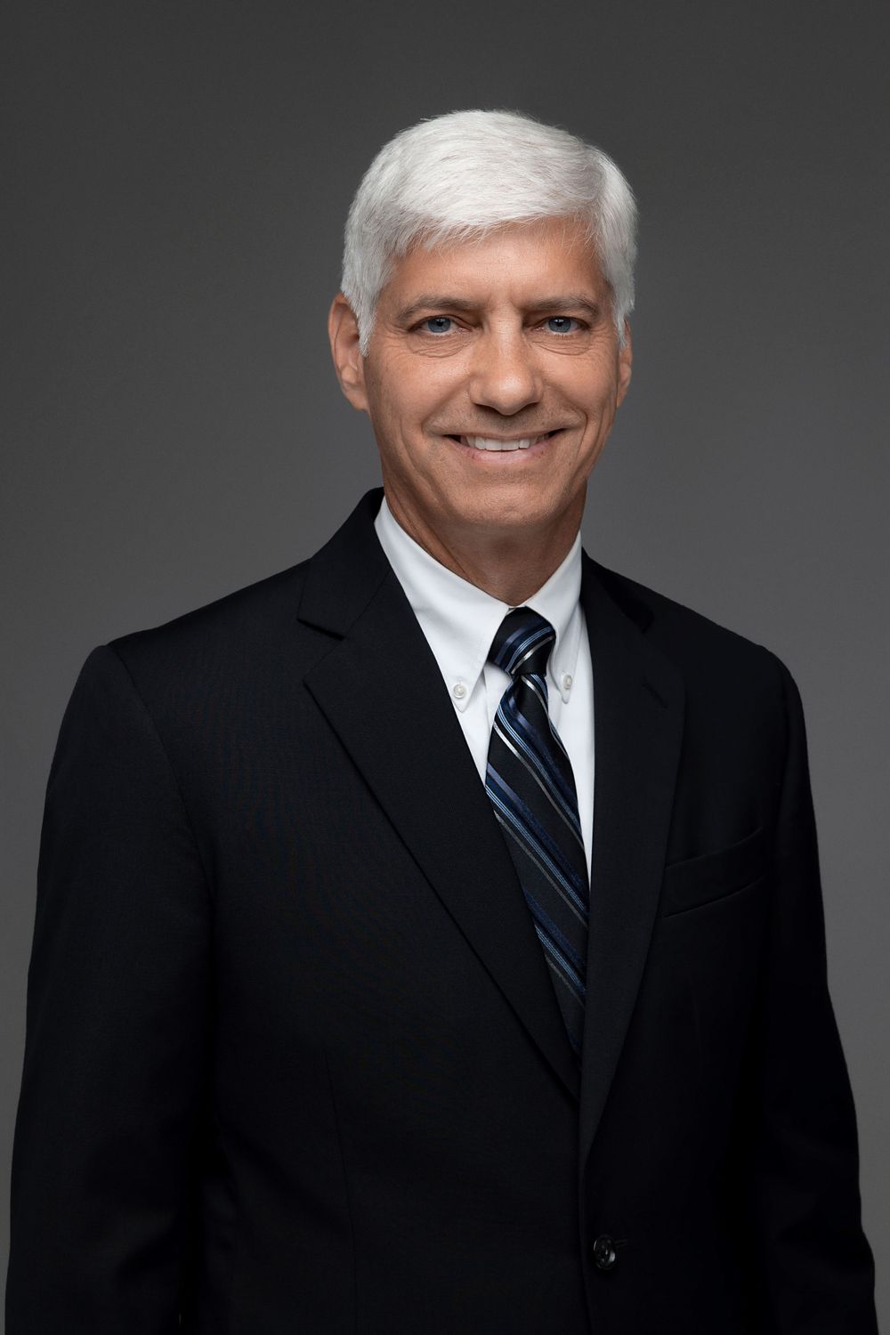 Executive portrait of person in dark suit with striped tie against grey background.