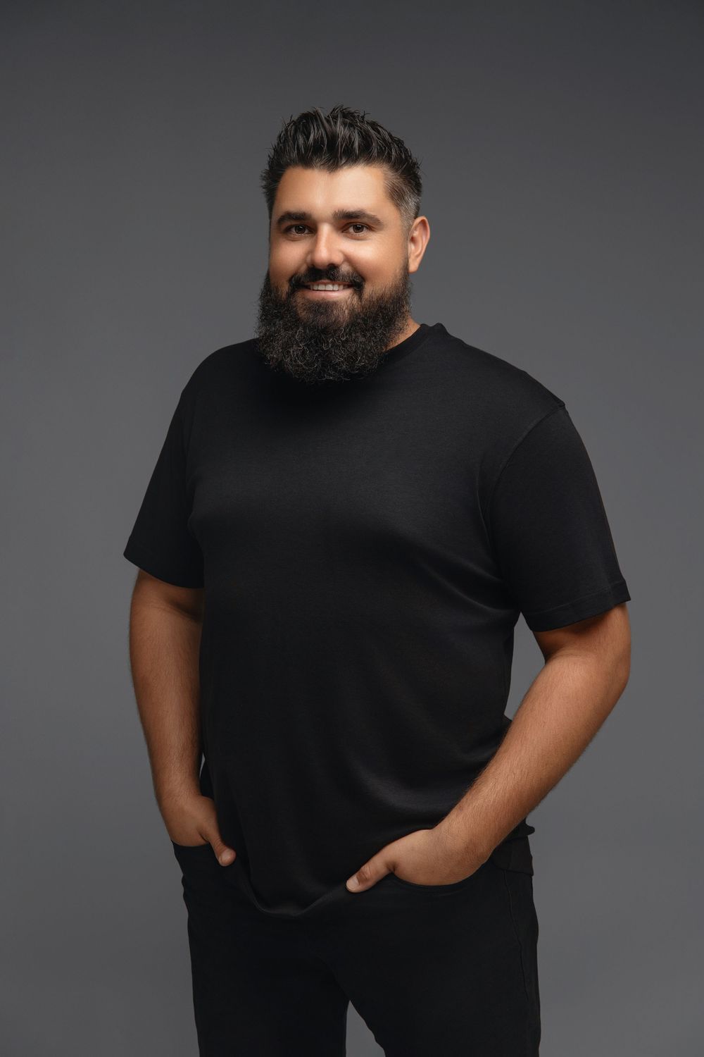 Casual portrait of bearded man in black t-shirt with hands in pockets against dark backdrop.