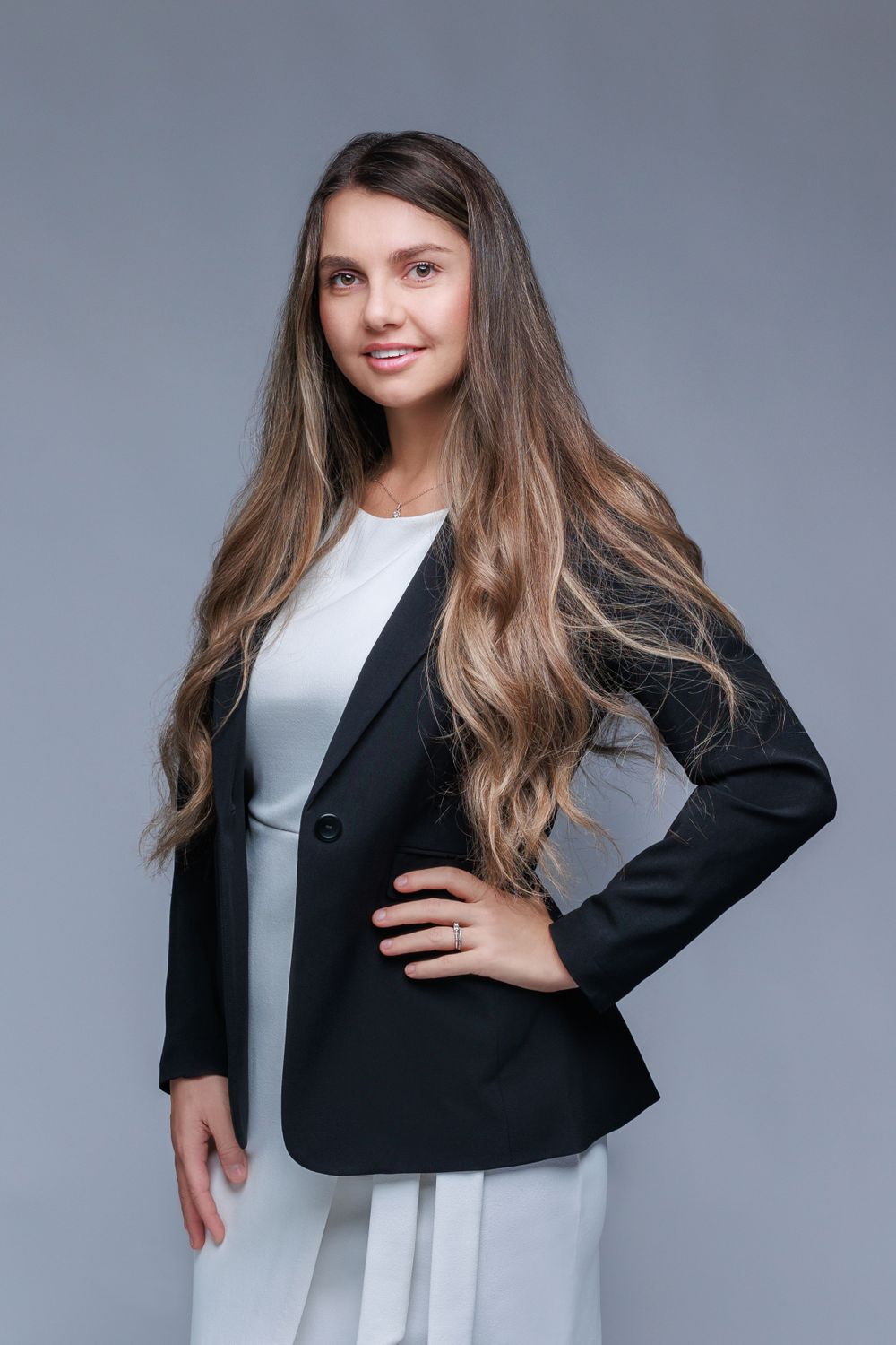 Professional portrait of smiling businesswoman in black blazer and white dress against gray backdrop.