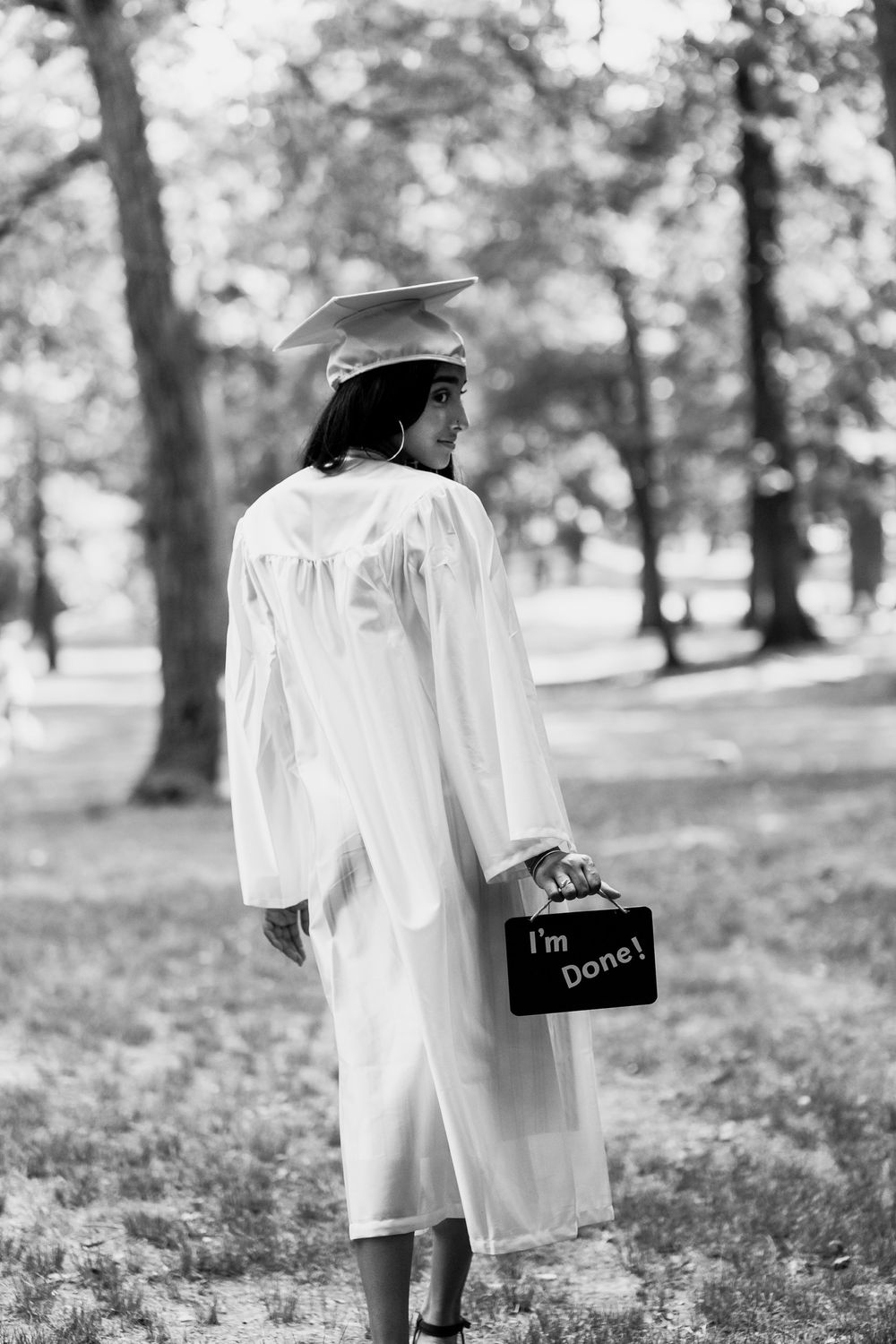 A graduate in white cap and gown walks away holding a sign that reads 'I'm Done' in a park setting.