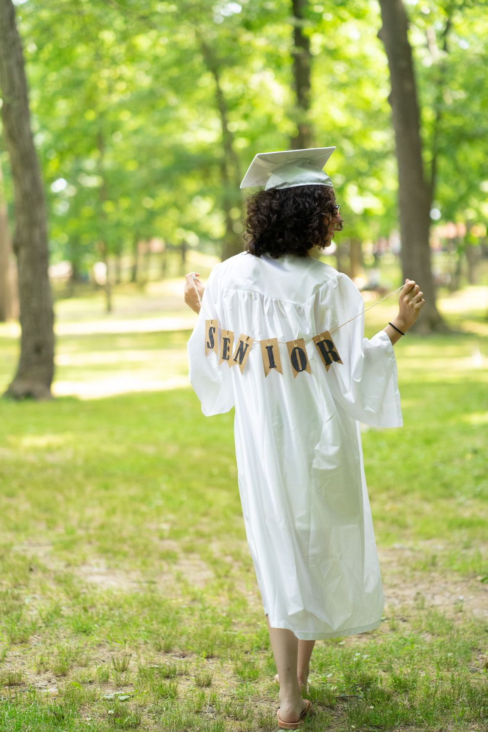 Graduate in white cap and gown walks outdoors holding a banner displaying 'Senior' text in a sunny woodland setting.