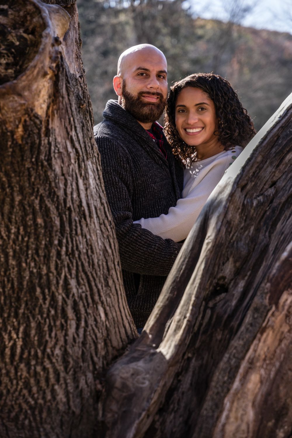 Couple poses intimately between tree trunks during outdoor portrait session.