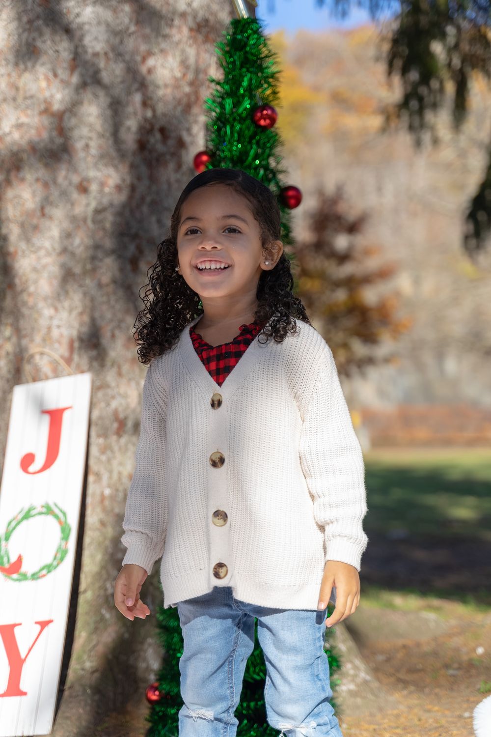 Child wearing white cardigan poses with Christmas decorations and JOY sign outdoors.