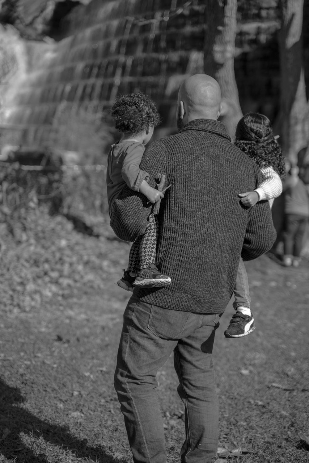 Back view of person carrying two children while looking at waterfall in black and white.