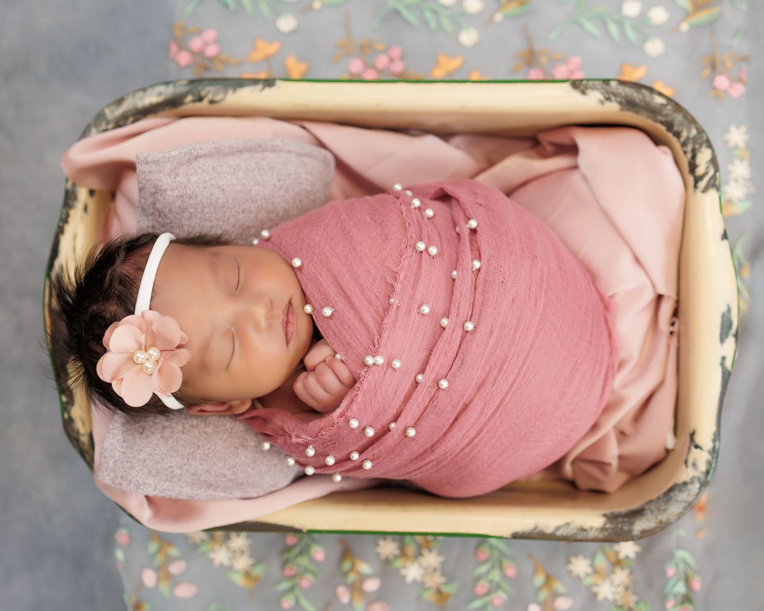 A sleeping newborn baby wearing a pink knitted outfit rests peacefully in a bassinet with soft pink bedding.
