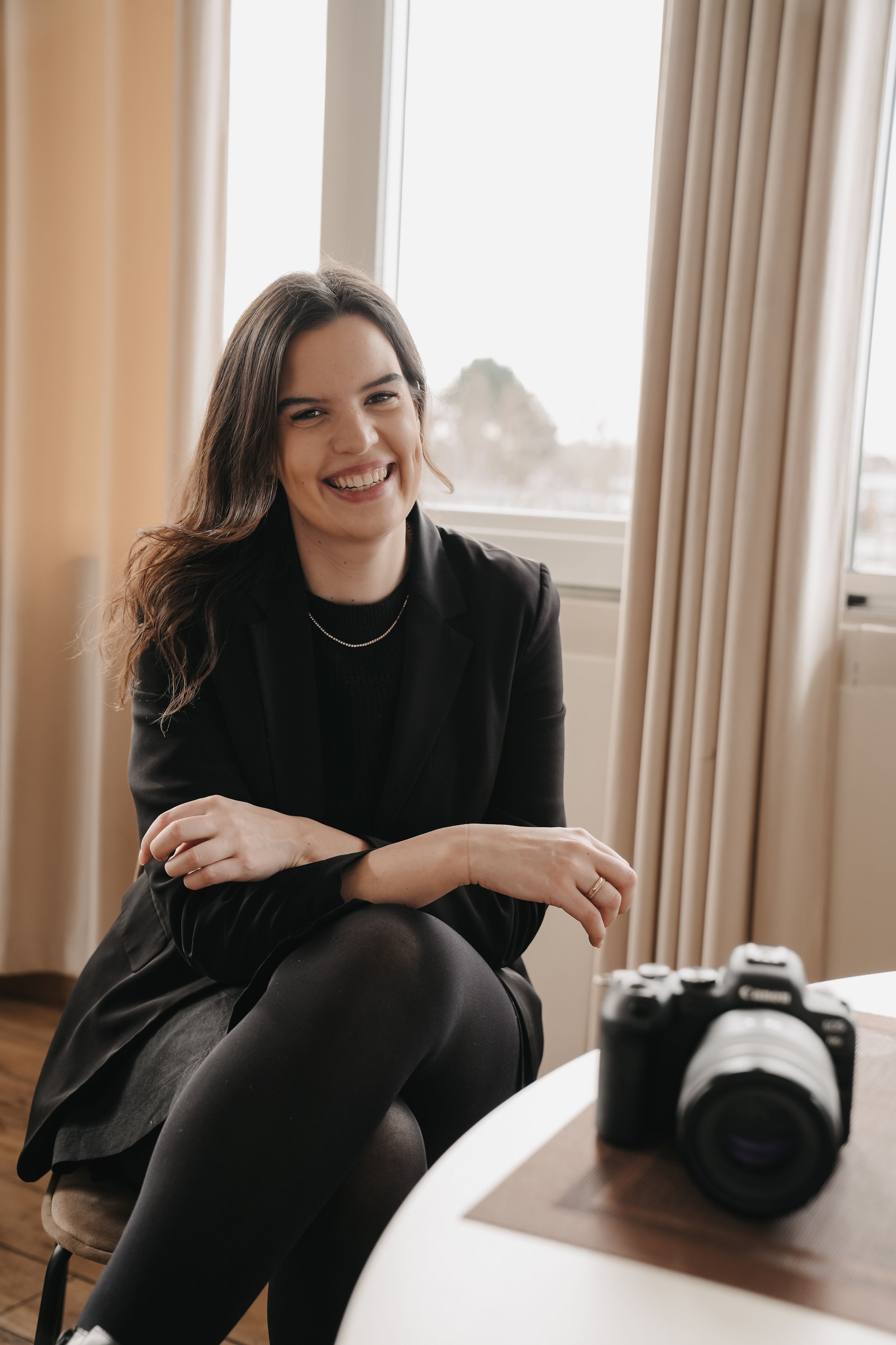 A photographer sits casually on a chair next to their camera in a warmly lit room with curtained windows.